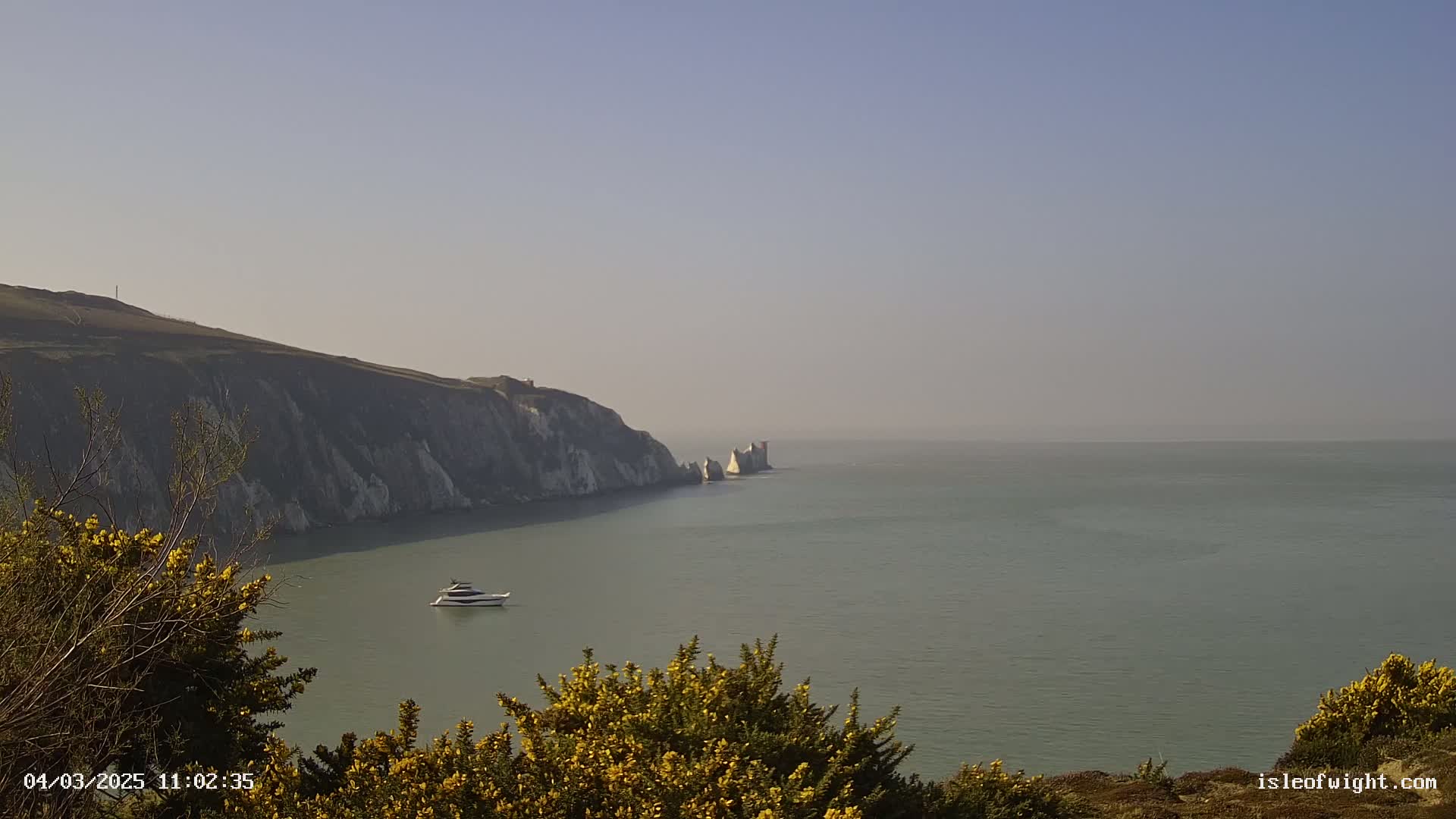 A hazy, calm sea stretches out before a white cliff face, with a lone boat visible in the distance.