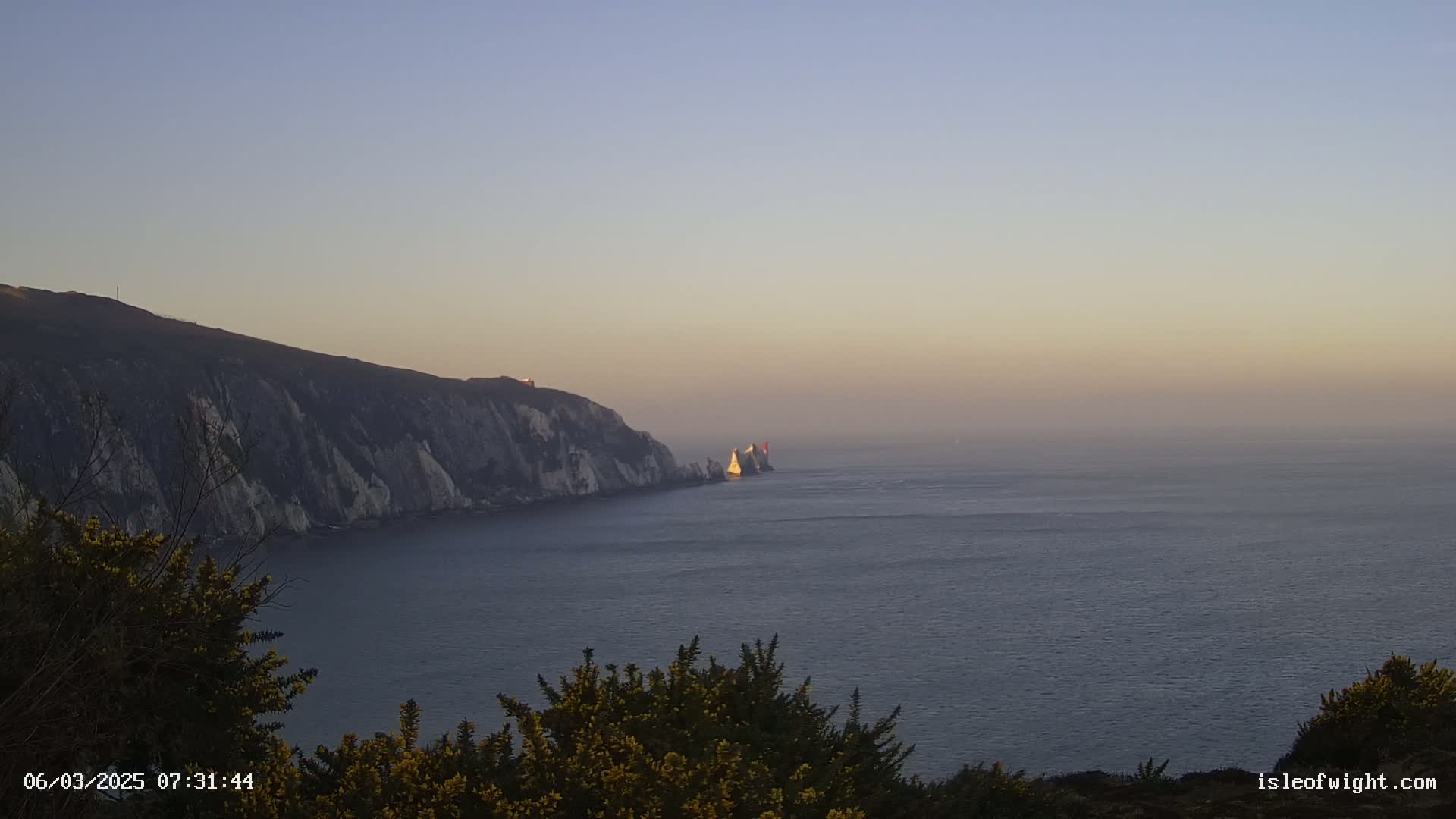 White cliffs and a rocky outcrop are visible across a calm sea under a hazy sunset.