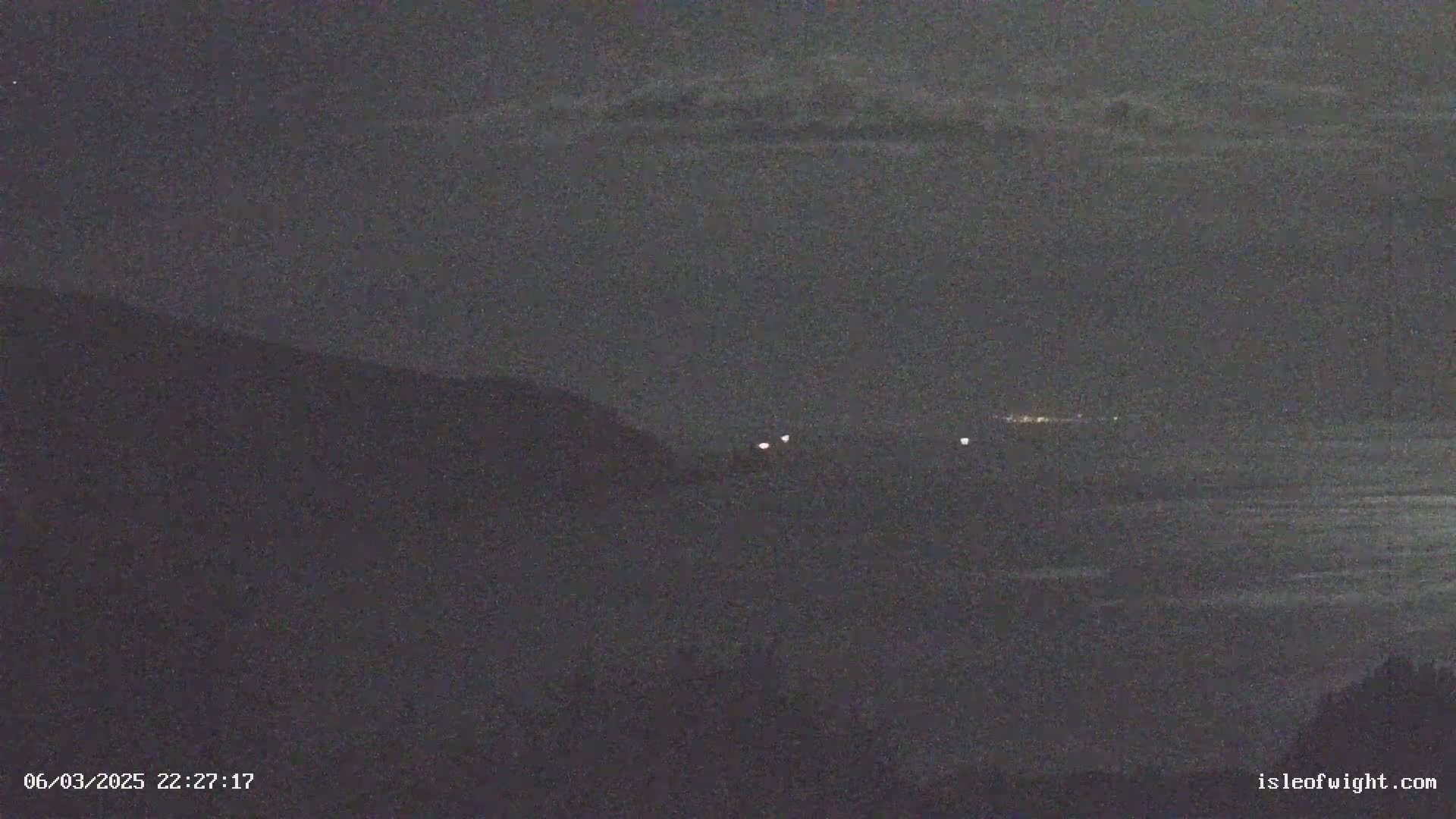 A nighttime, dark landscape shows a hill overlooking a body of water with a few distant lights visible under a cloudy sky.