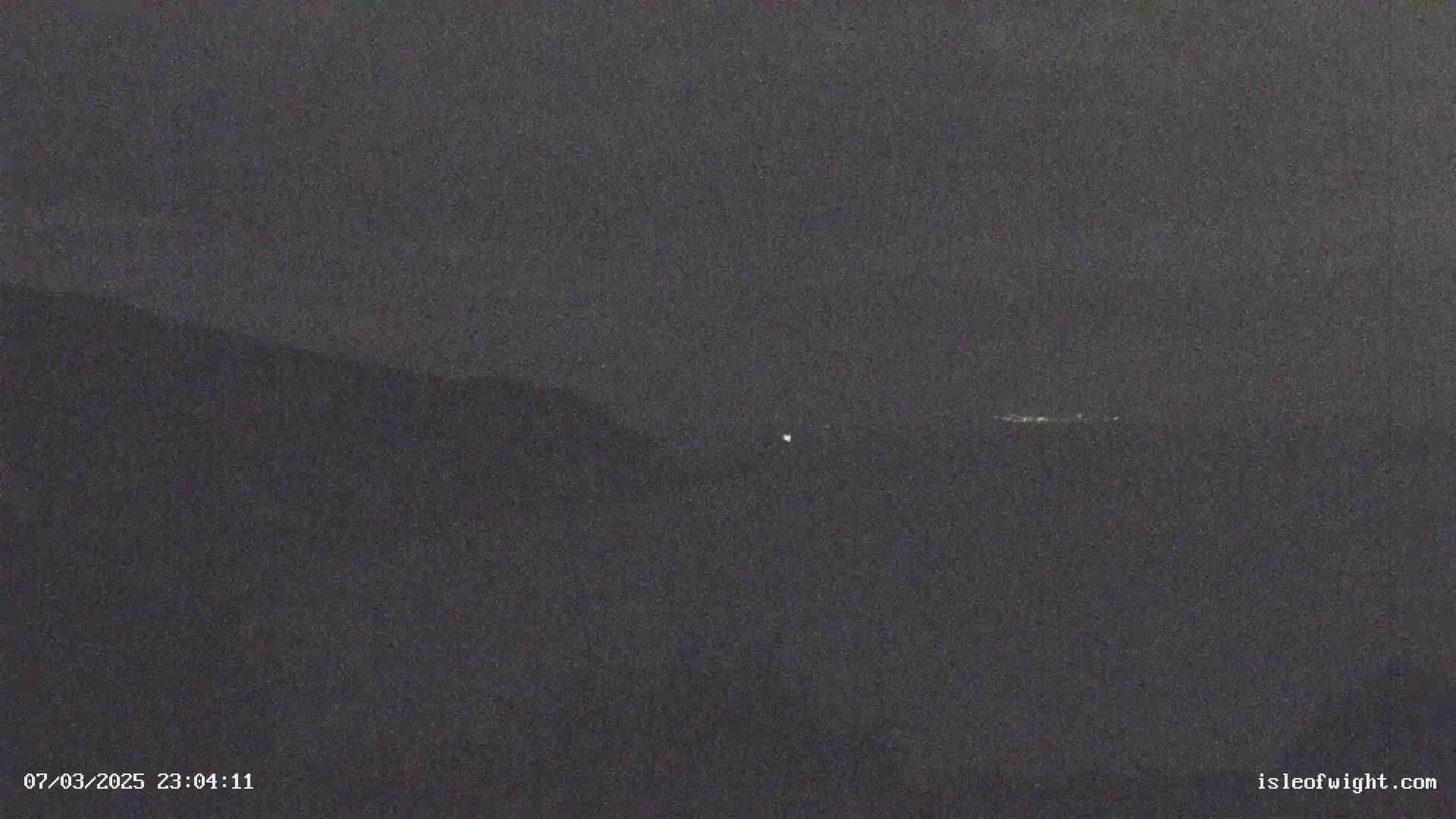 A dark, nighttime view of distant lights and hills under a cloudy sky.