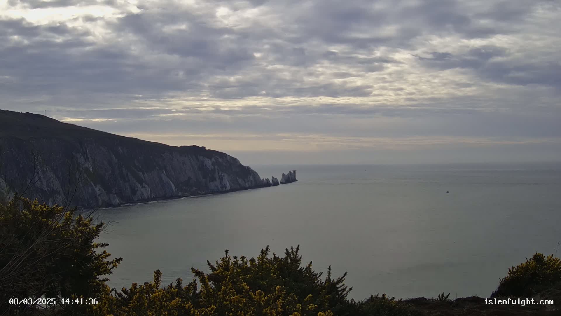 A coastal scene under a mostly cloudy sky shows a steep, light-colored cliff face extending into a calm sea, where a small cluster of rocks is visible further out.
