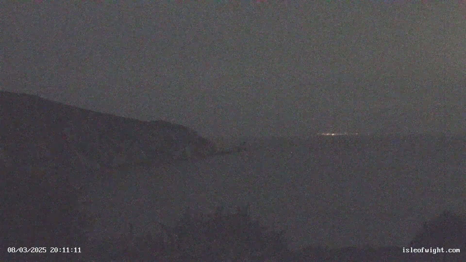 A dark, nighttime view shows a distant line of lights across a hazy horizon, with a dark hill or cliff in the foreground.