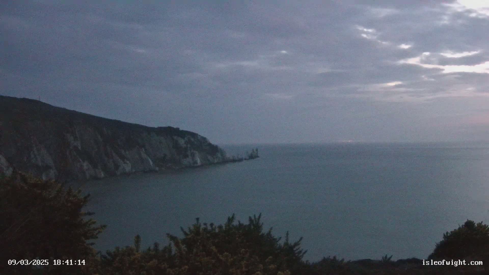 A dark, overcast sky hangs over a calm sea, with a cliff face and dark shrubbery visible in the foreground.