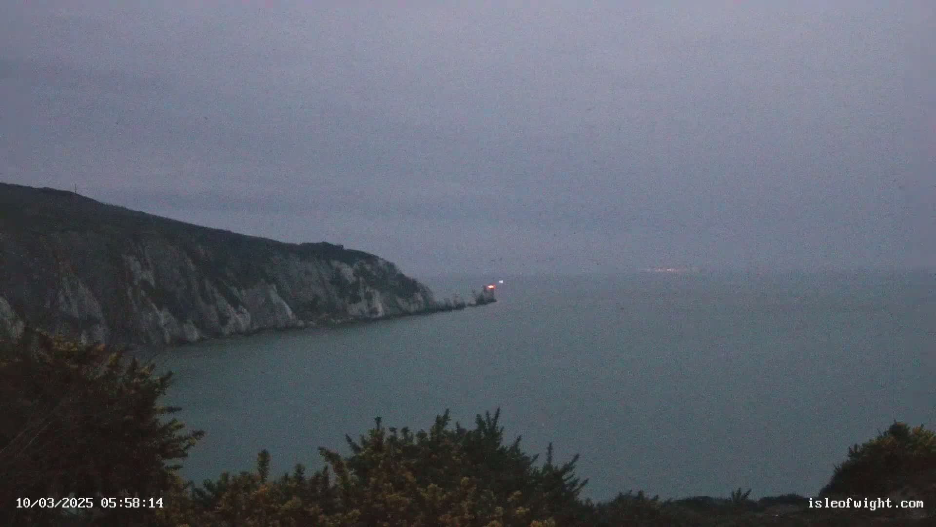 A lighthouse sits on a rocky outcropping near a large, cliff-edged landmass, all under a hazy, overcast sky.