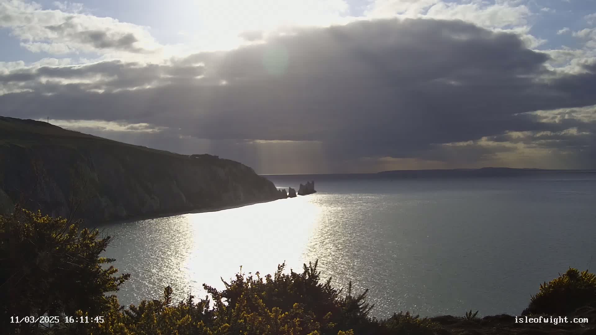 A partly sunny day shows a rocky coastline and calm sea reflecting sunlight, with dark clouds overhead.