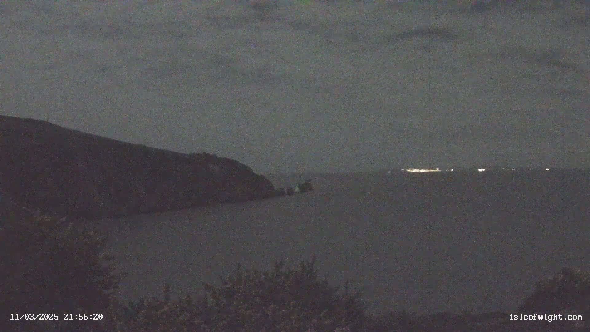 A dark, nighttime scene shows a body of water with a distant shoreline, illuminated by faint lights, and a silhouetted hill or mountain in the foreground under a cloudy sky.