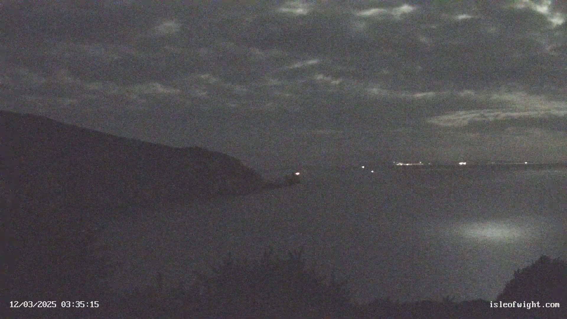 A nighttime, cloudy view shows a dark hillside overlooking a body of water with distant lights on the horizon.
