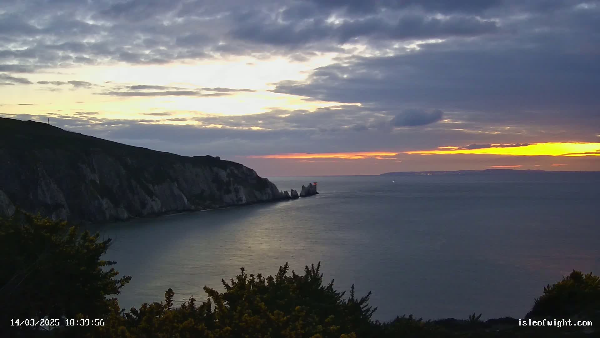 A sunset over a calm sea with a cliff face and a small island visible in the distance under a partly cloudy sky.