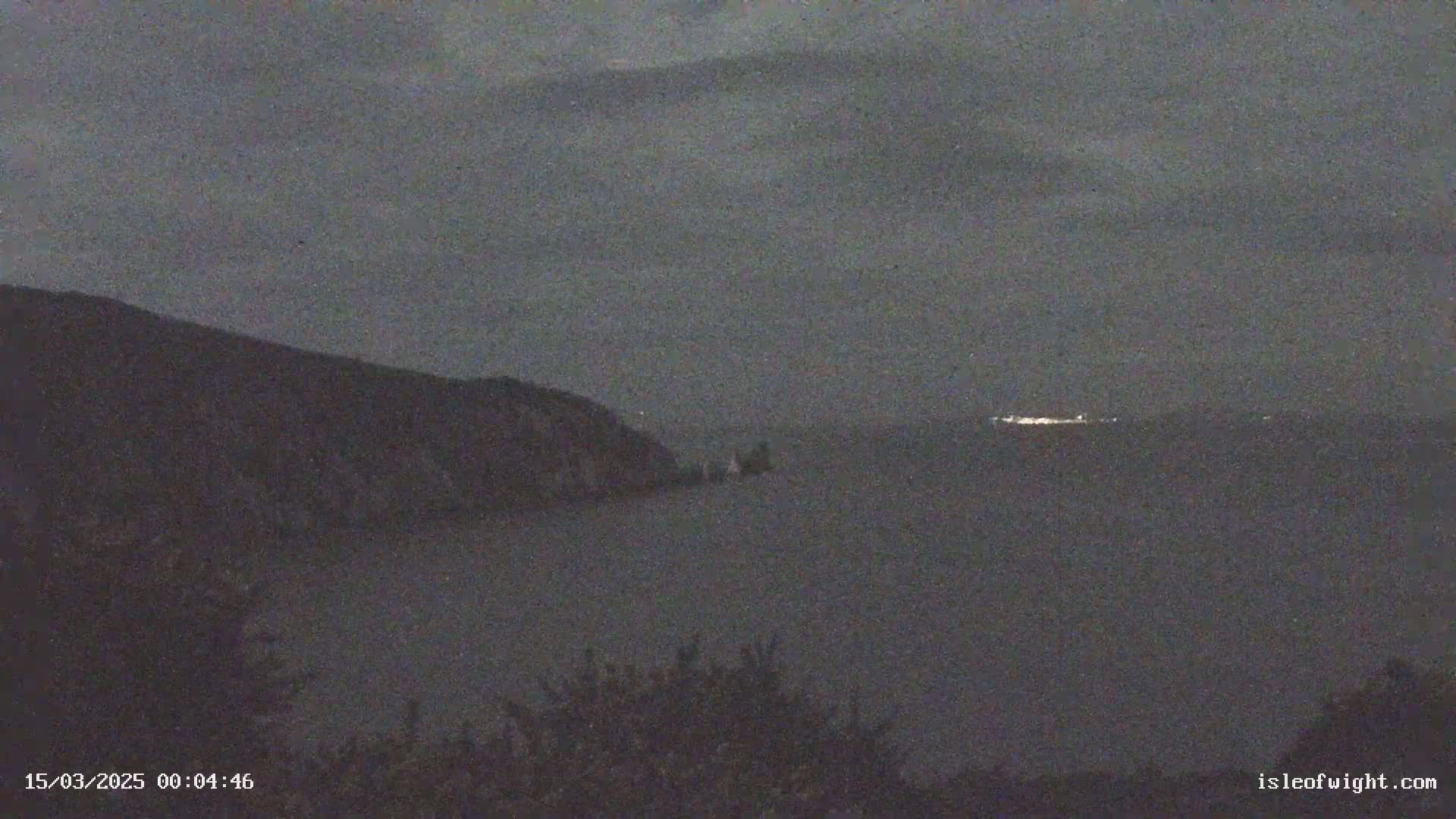 A dark, overcast night scene shows a body of water with a distant shoreline faintly illuminated, and a dark hillside in the foreground.