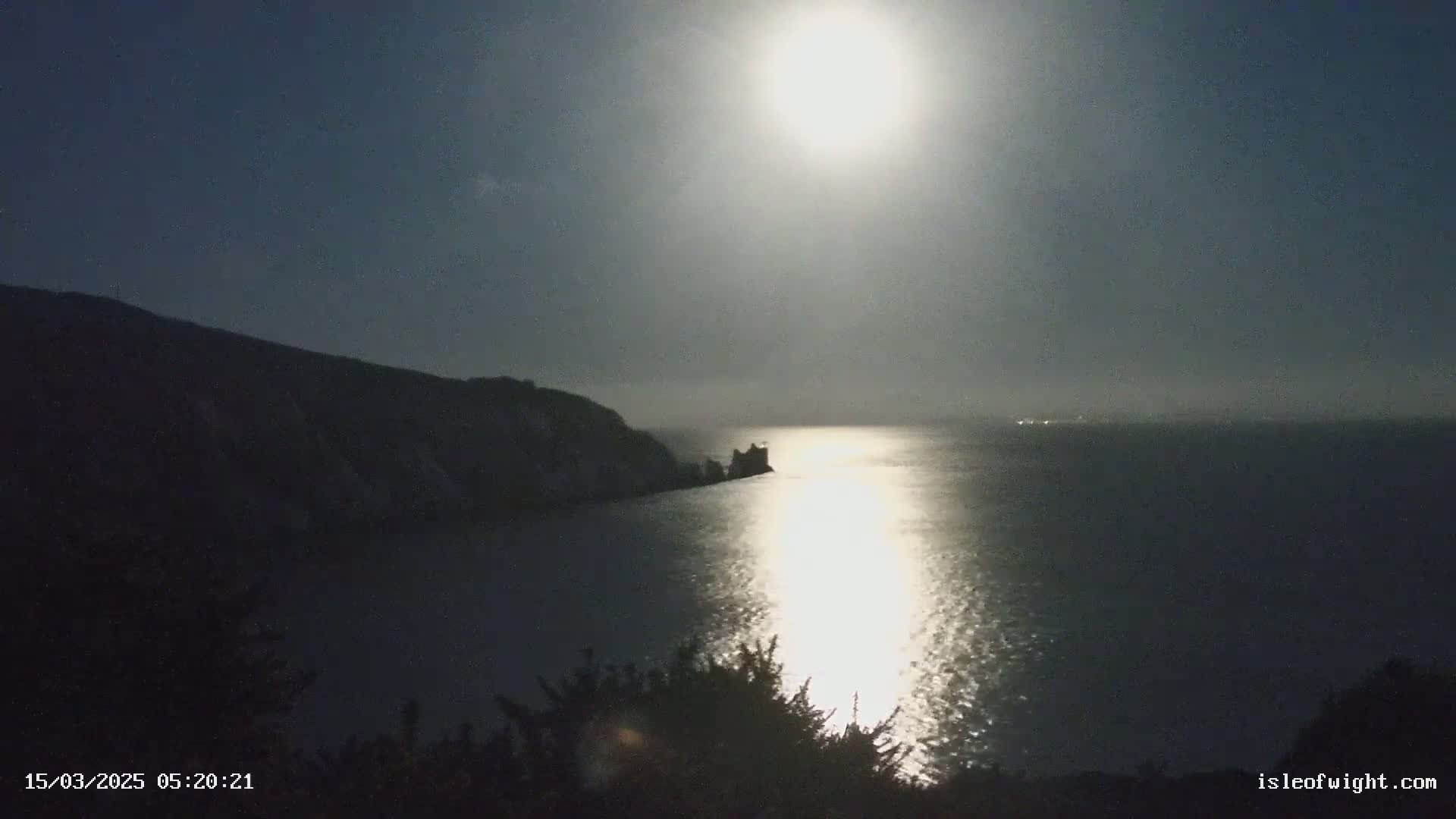 A bright moon illuminates a calm body of water at night, reflecting its light on the surface near a dark, rocky coastline.