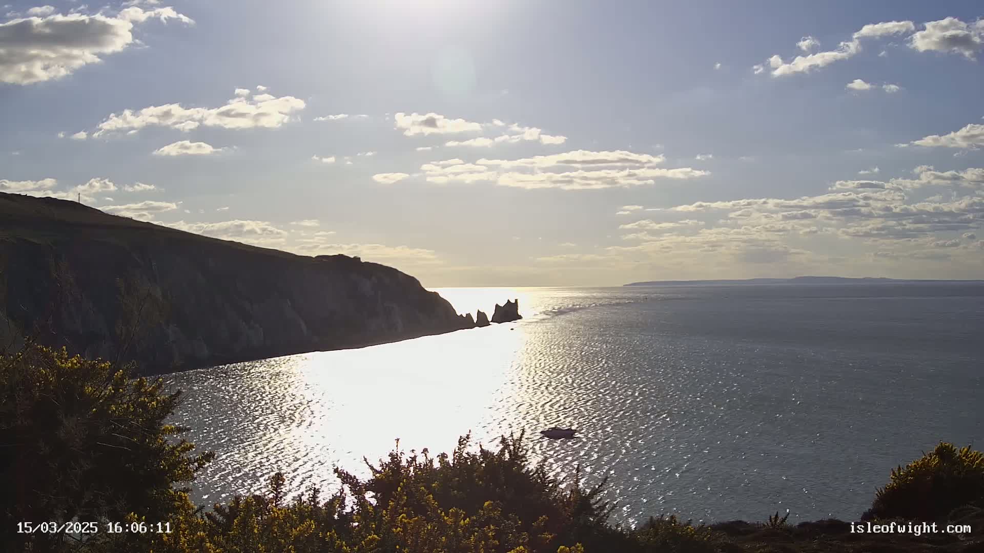 A sunny day with partly cloudy skies shows a view of a calm sea reflecting sunlight, with a cliff and some rock formations in the foreground.