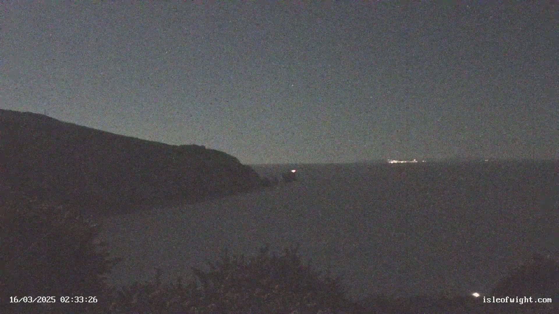 A dark, nighttime coastal scene shows a silhouetted hill overlooking a calm sea with distant lights and a faint fog or mist.
