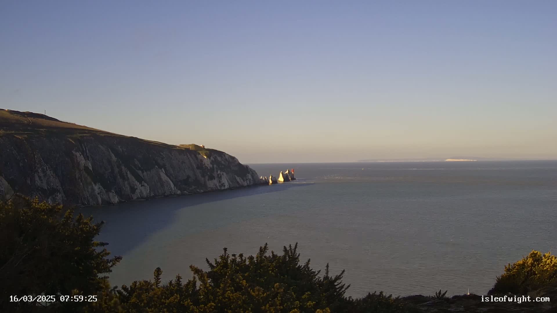 A coastal scene shows a cliffside, the ocean, and distant land under a clear, light blue sky.