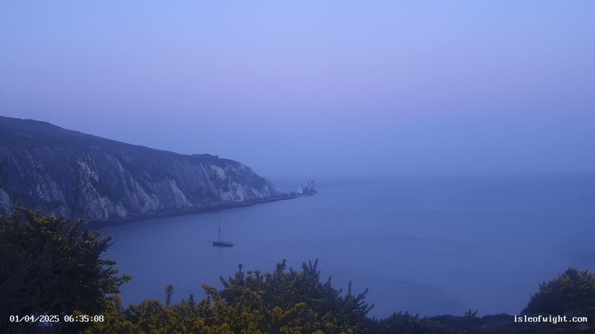 A hazy, blue-toned view from a high vantage point shows a sailboat on calm water next to white cliffs.
