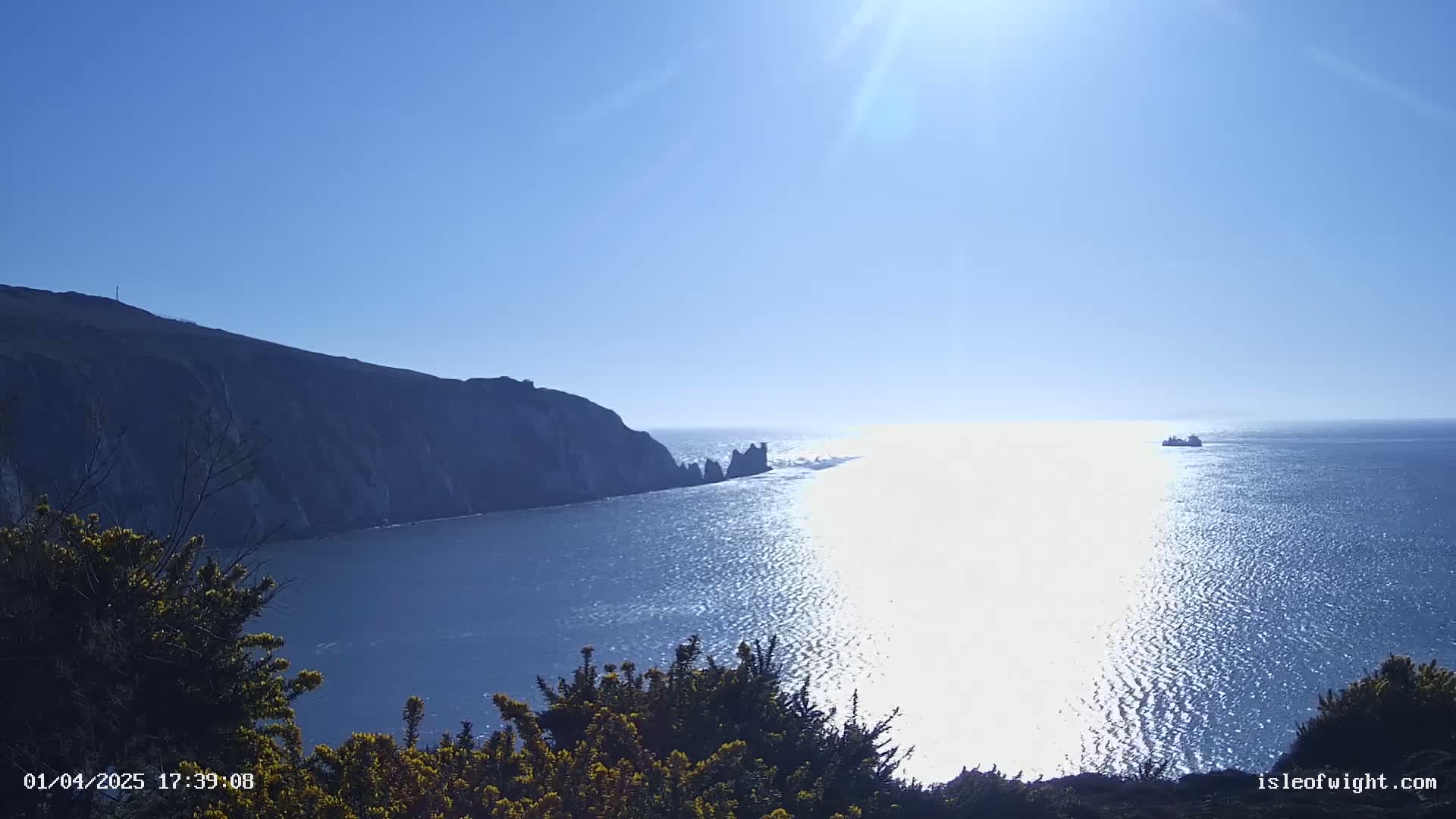 A sunny day reveals a coastal scene with dark green cliffs meeting a bright blue ocean, where a ship is visible on the horizon.