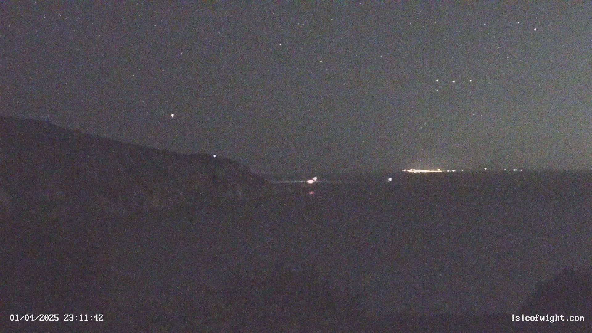 A nighttime, starlit view shows a dark landmass in the foreground and a faint, distant line of lights on the horizon across a dark body of water.