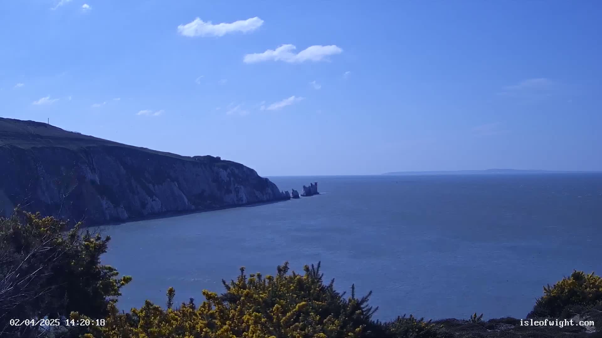 A partly cloudy blue sky overlooks a calm sea, where a white cliff and several smaller rock formations jut out from a green hillside.