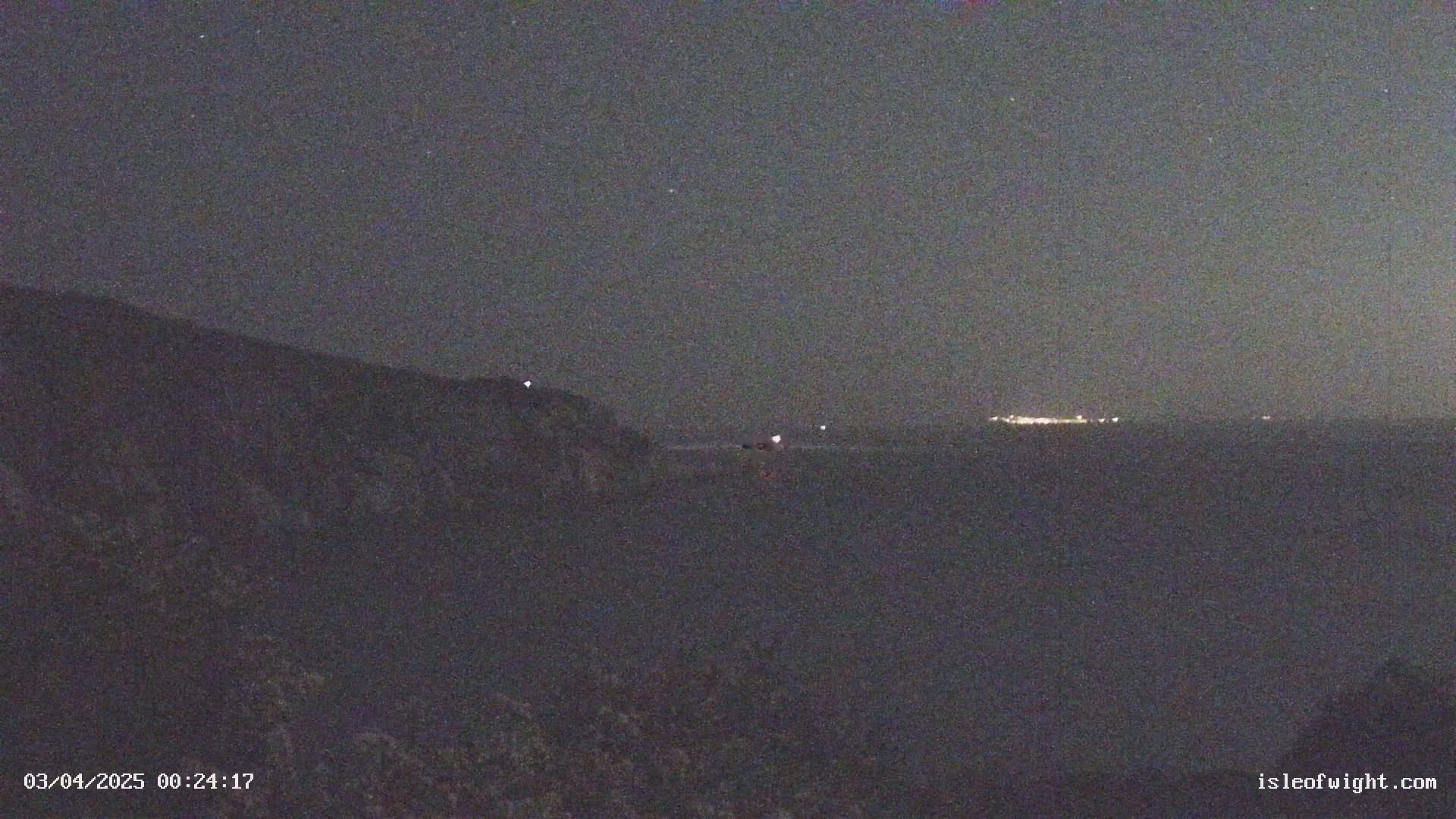 A dark, nighttime scene shows a body of water with a distant, faintly lit shoreline, and a dark hillside in the foreground under a clear, starry sky.