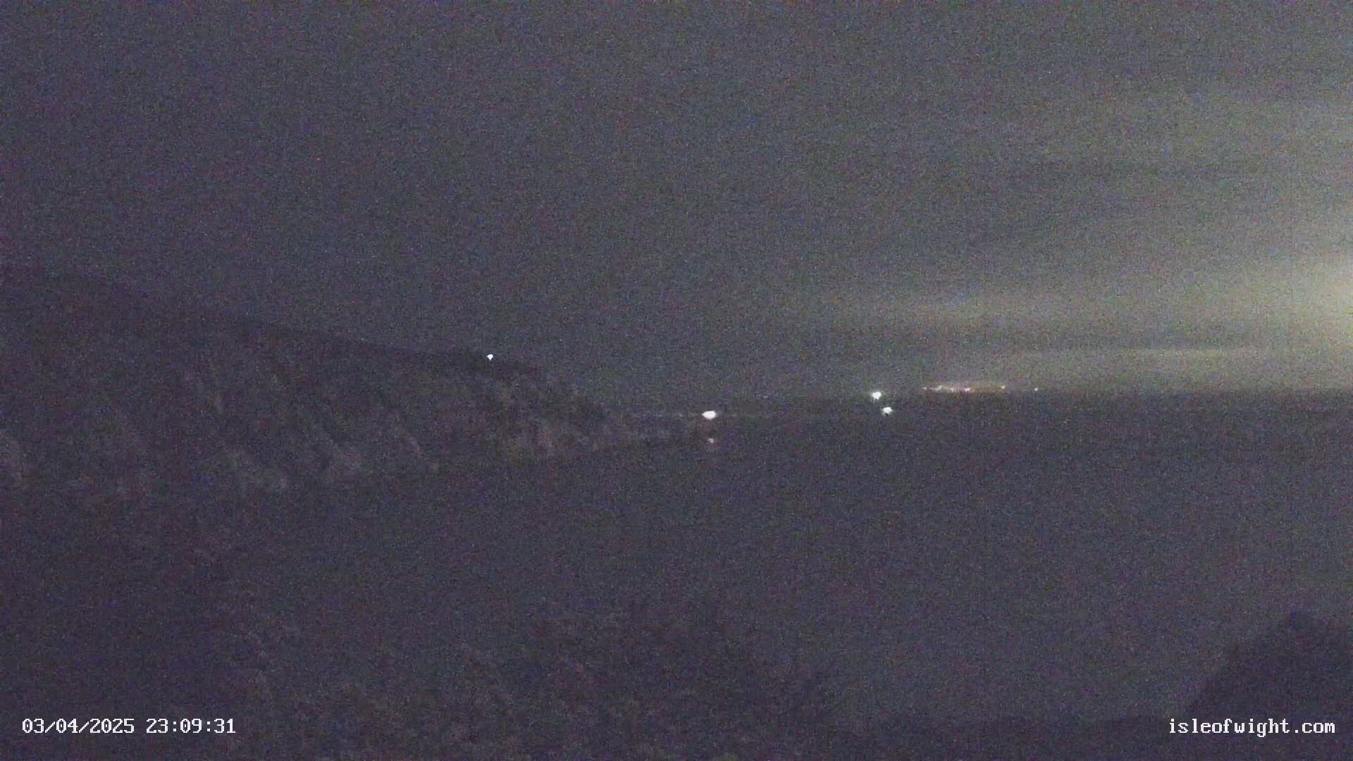 A nighttime, long shot of a dark landscape with a few distant lights visible on a horizon line under a mostly clear, dark sky.
