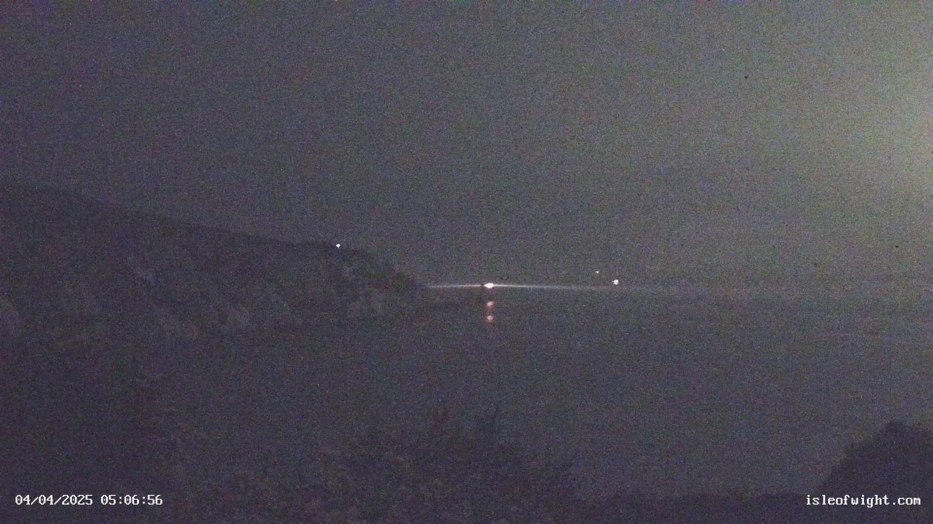 A dark, nighttime scene shows a body of water with faint lights in the distance, possibly from ships, and a dark landmass in the foreground under a cloudy or hazy sky.