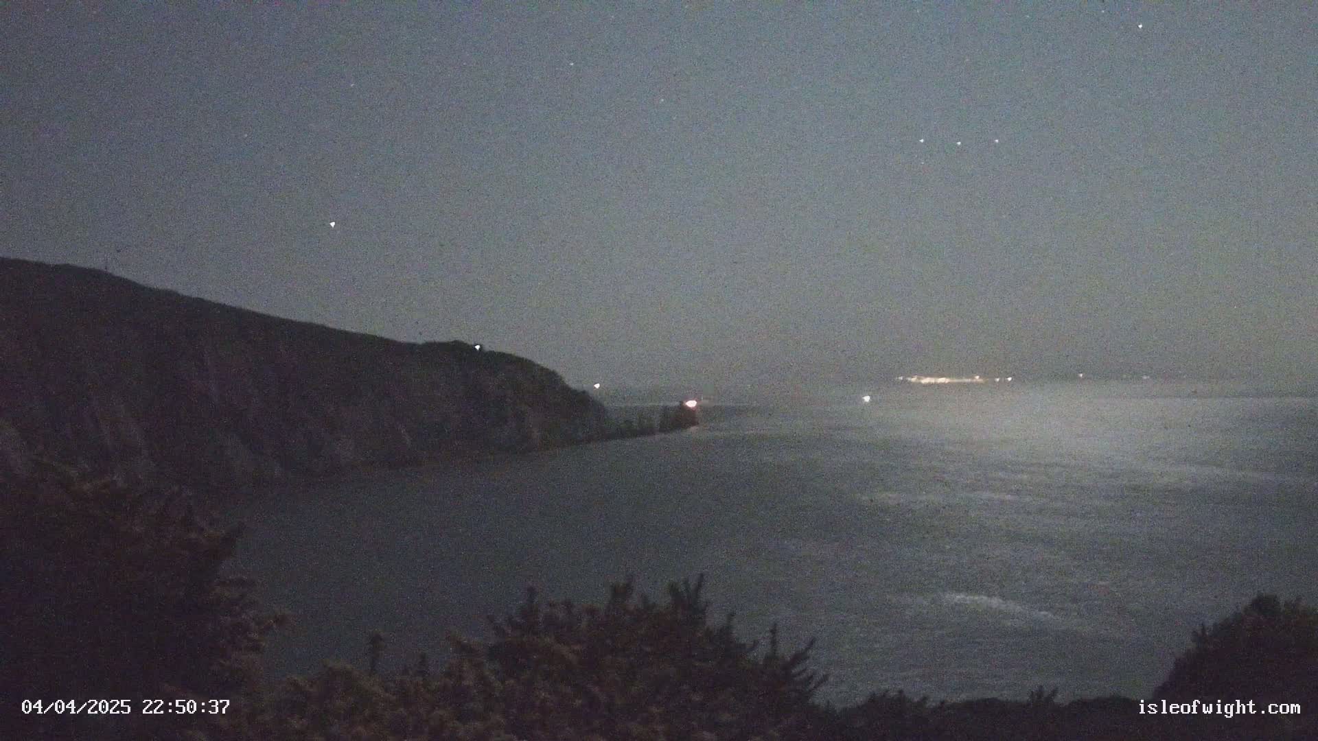 A dark, nighttime coastal scene shows a cliff overlooking a calm body of water with faint lights visible in the distance under a mostly clear, starry sky.
