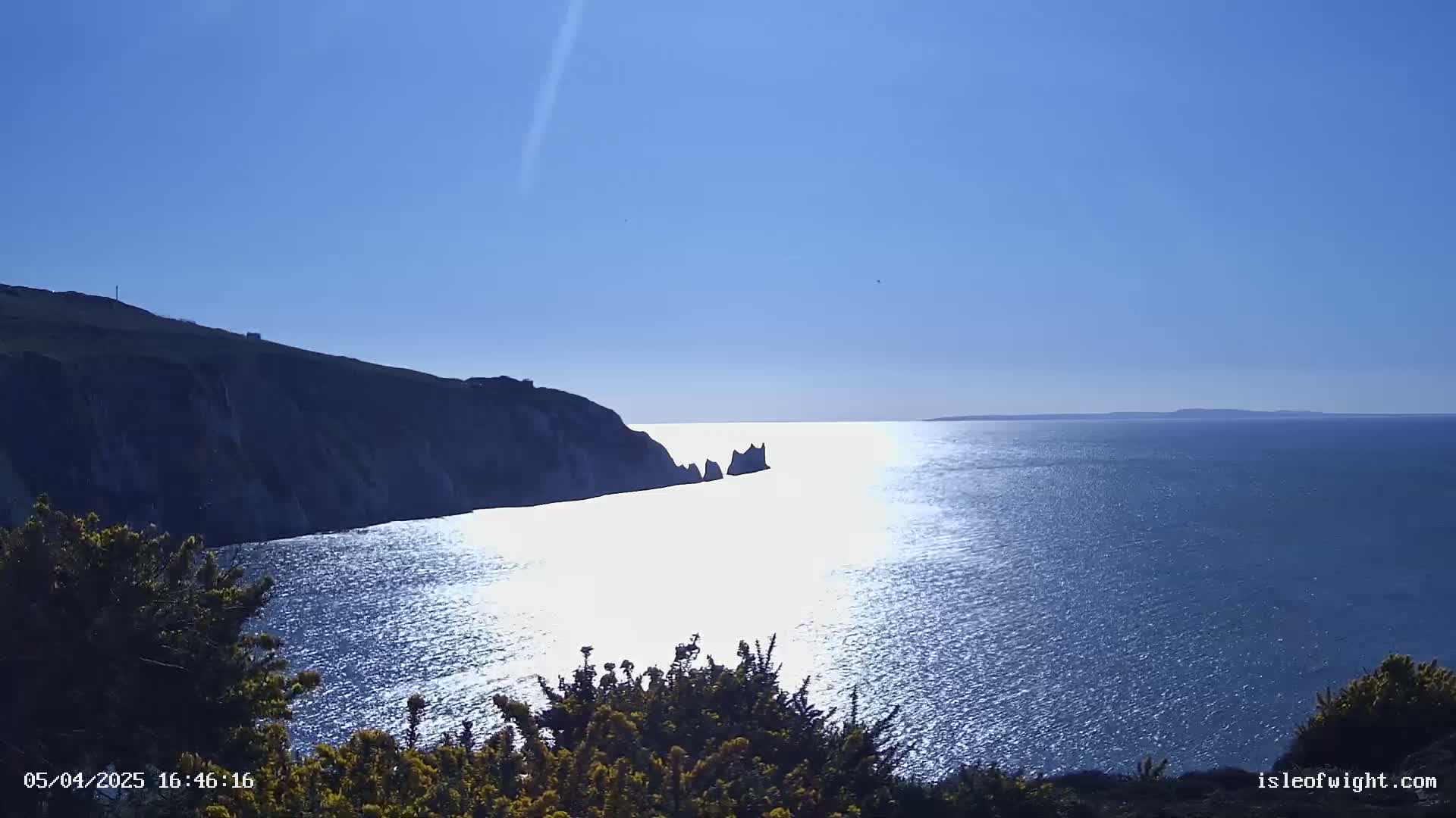 A sunny day reveals a scenic coastal view of a dark cliff overlooking a calm, sunlit ocean with a distant, small island or rock formation.