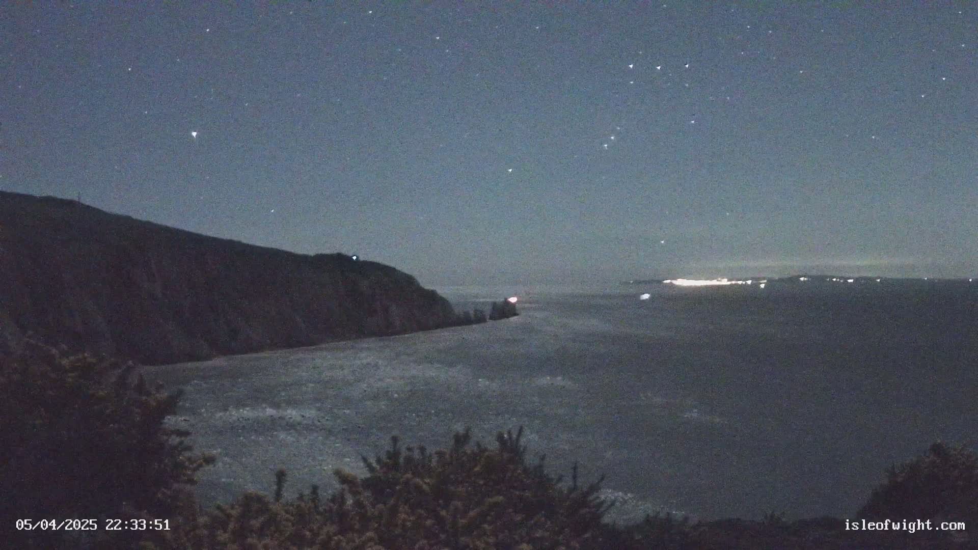 A dark, rocky cliff overlooks a calm, dark body of water under a clear, starry night sky.