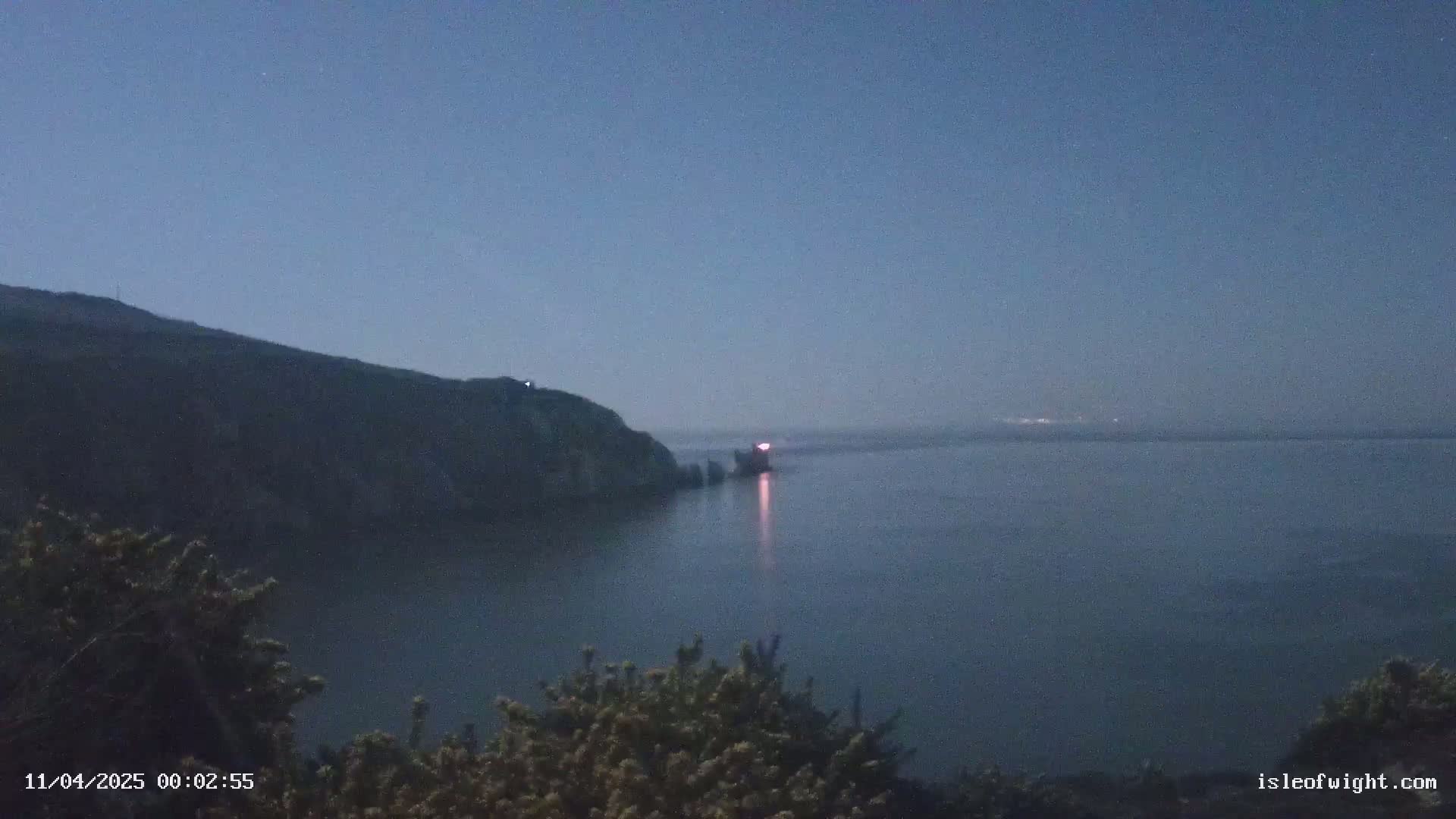 A dark, calm body of water is seen at twilight, with a rocky headland and a distant light source on the opposite shore.