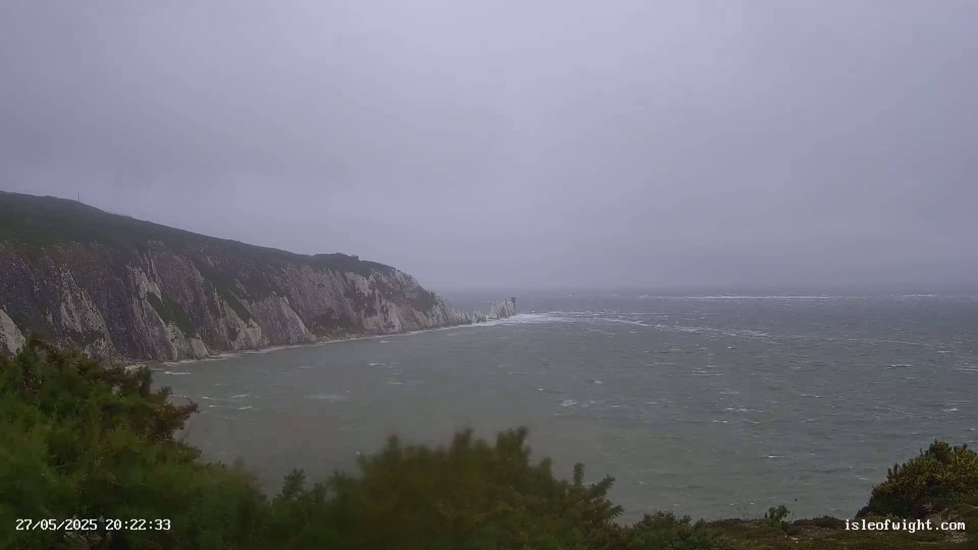 A view from a clifftop shows a windy, overcast day over a choppy sea with white cliffs extending into the distance.