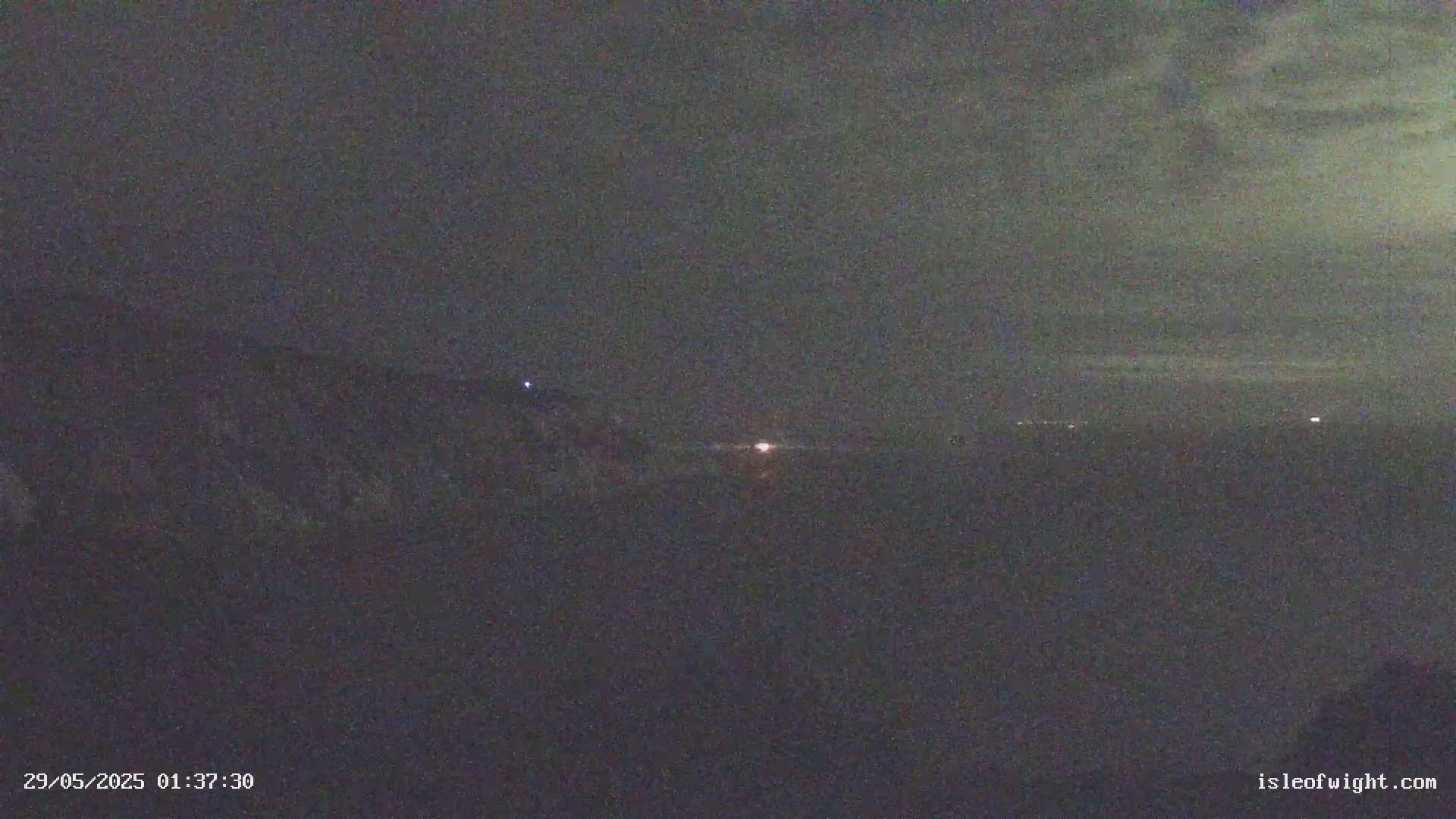 A nighttime, low-light image shows a dark landscape with a few faint lights in the distance under a mostly cloudy sky.