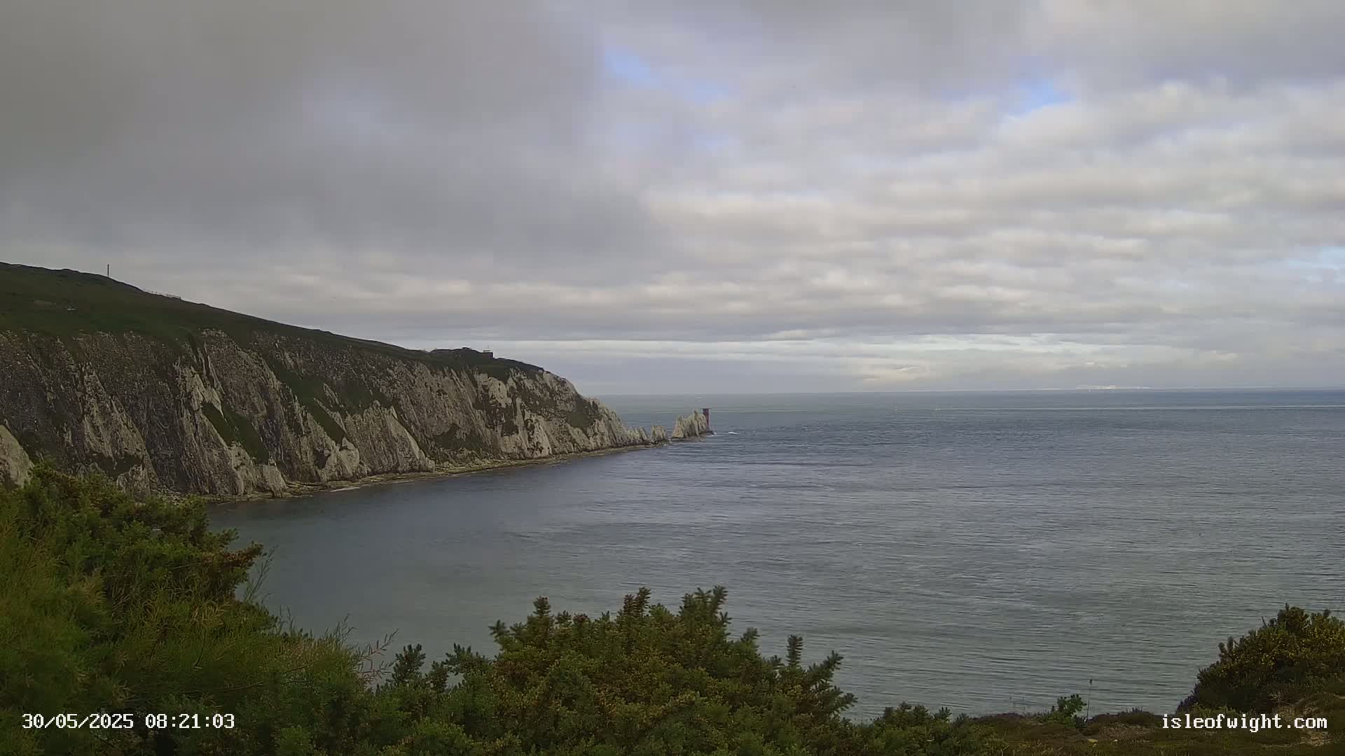 A coastal scene shows a rocky cliffside meeting a calm sea under a cloudy sky.