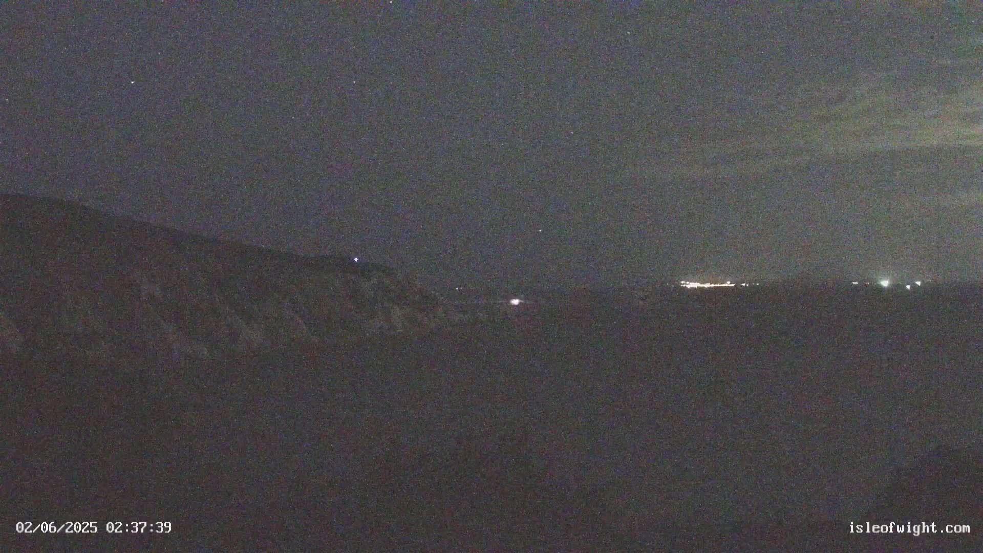 A nighttime view of a dark hillside overlooking a distant line of faint lights under a mostly clear, starry sky.