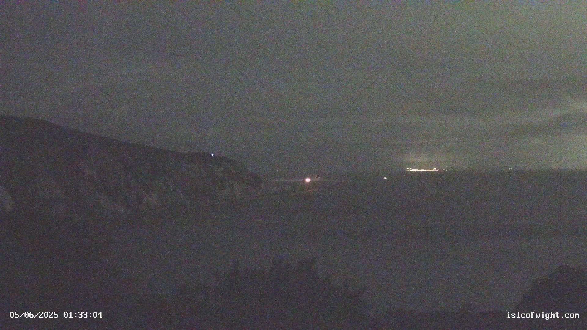 A nighttime, dark view shows a distant line of lights on the horizon behind a dark hill or mountain.