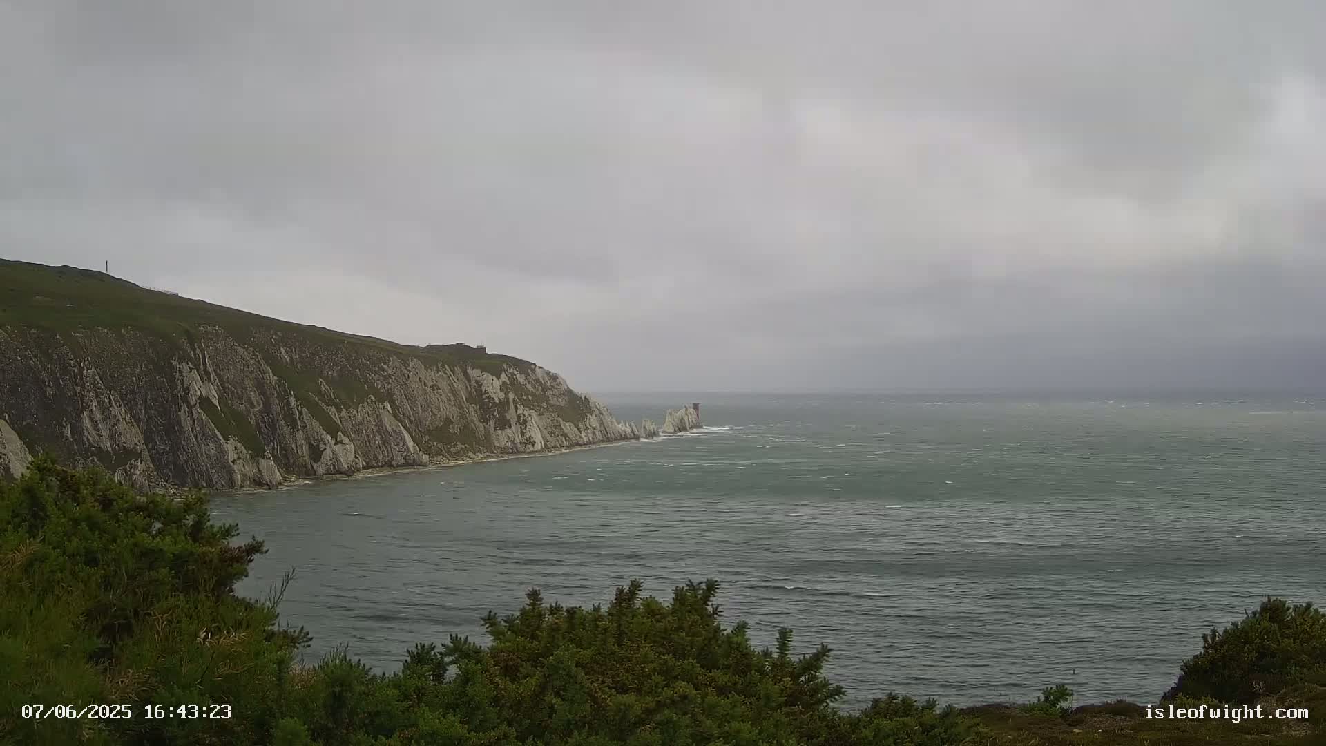 A windy, overcast day reveals a view of white cliffs meeting a choppy sea, with low-lying vegetation in the foreground.