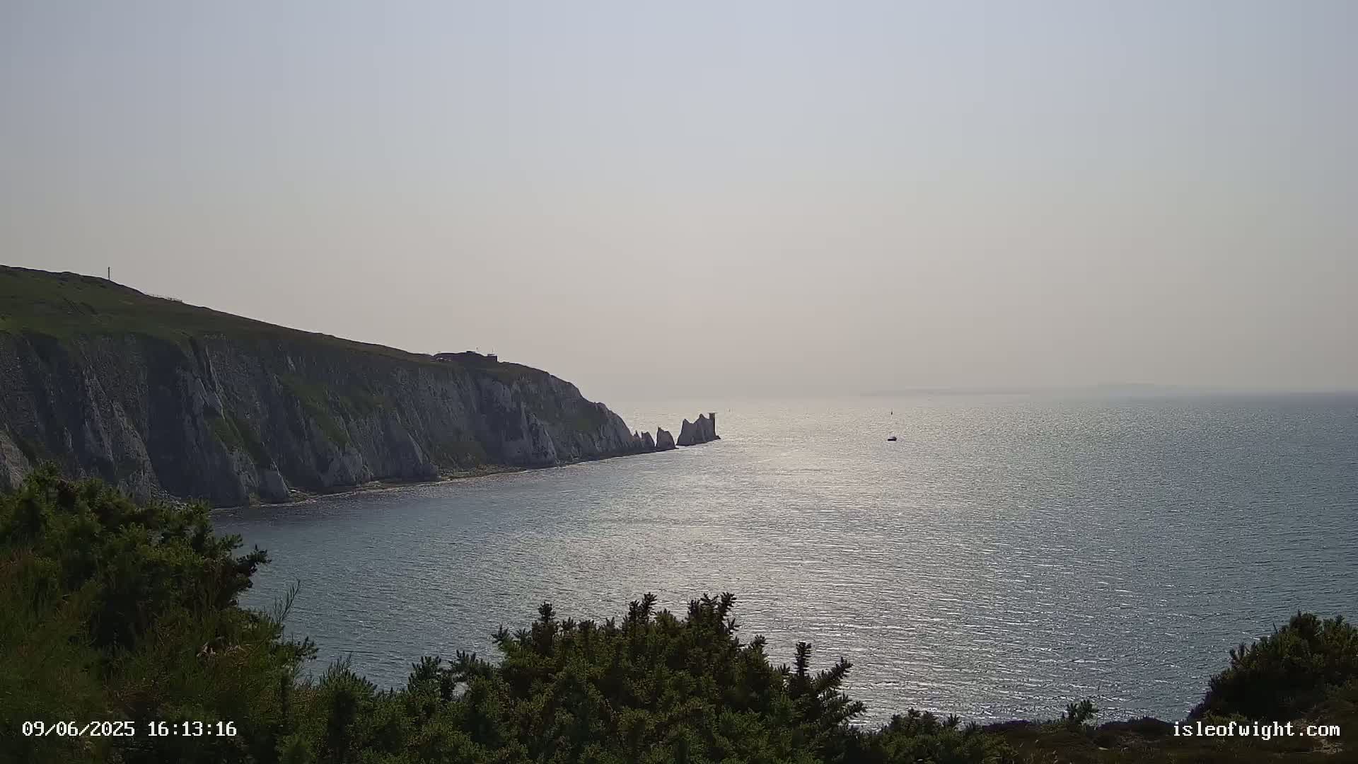 A hazy, sunny day reveals a view of a rocky coastline meeting a calm sea, with a small boat visible in the distance.