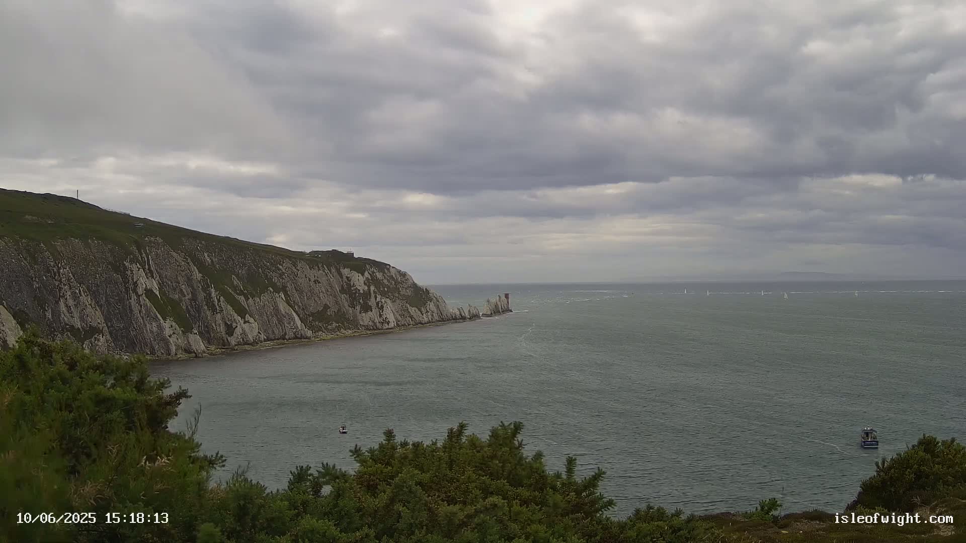 A coastal scene shows a gray, overcast sky over a calm sea with a few boats and a white cliff face extending into the water.