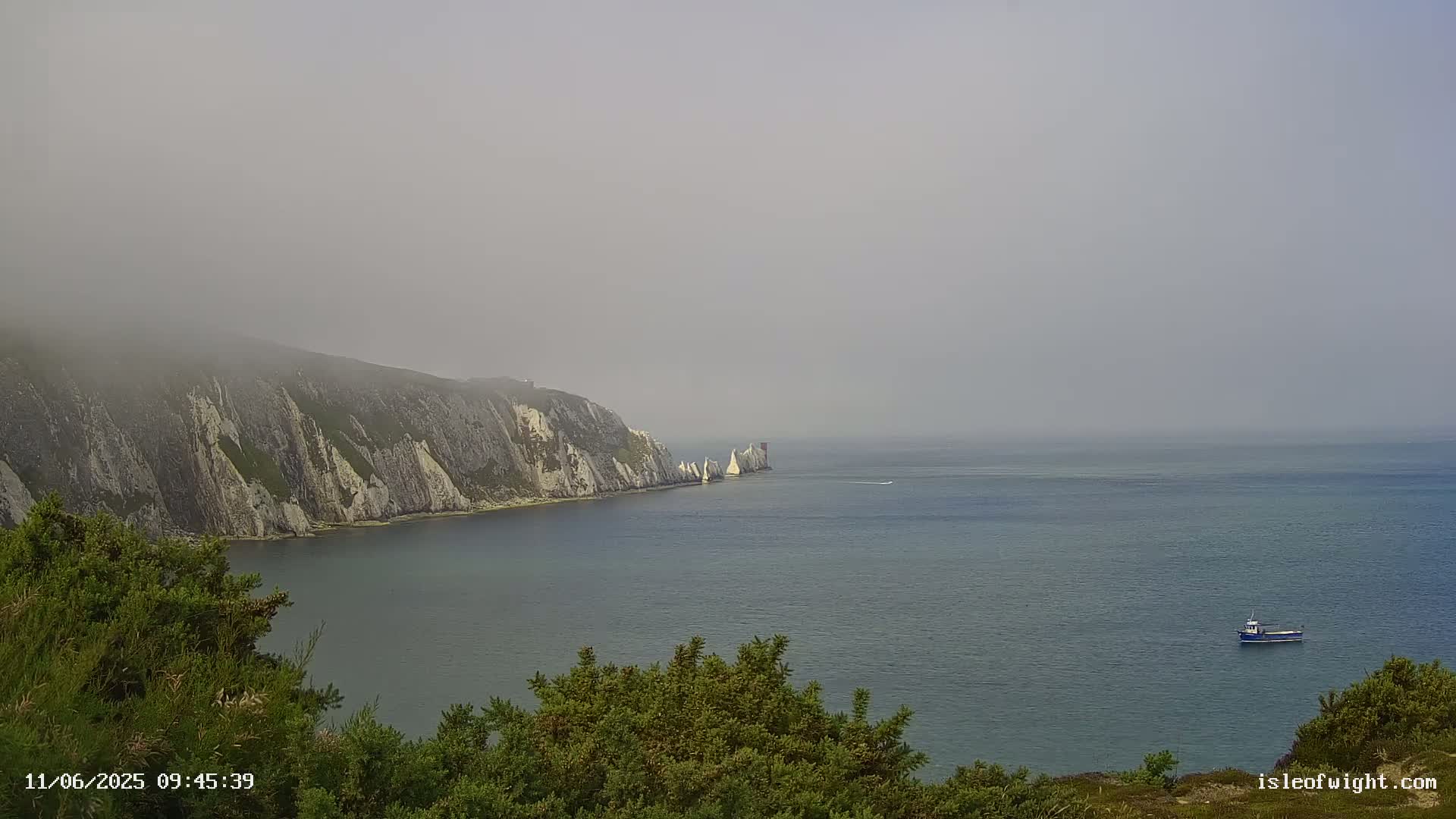 A hazy, foggy view of a rocky coastline with white cliffs and a small boat on the calm sea.