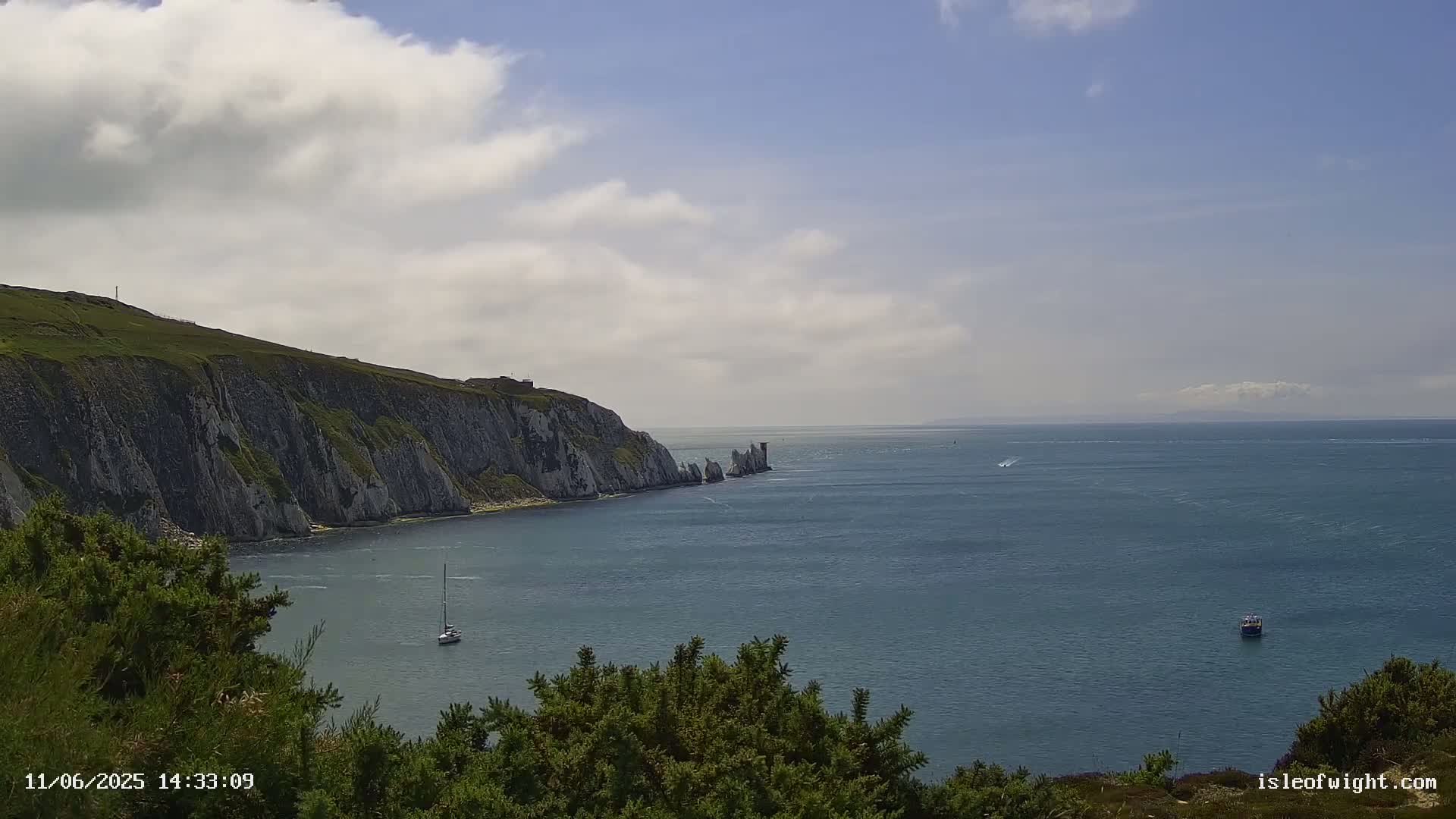 A partly cloudy day reveals a scenic coastal view of white cliffs, a sailboat, and a small boat on calm blue water.