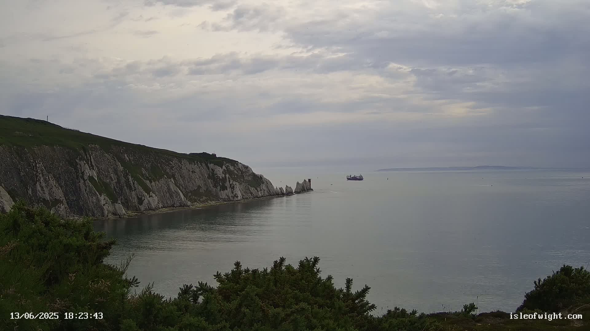 White cliffs meet a calm, gray sea under a cloudy sky, with a large vessel visible in the distance.