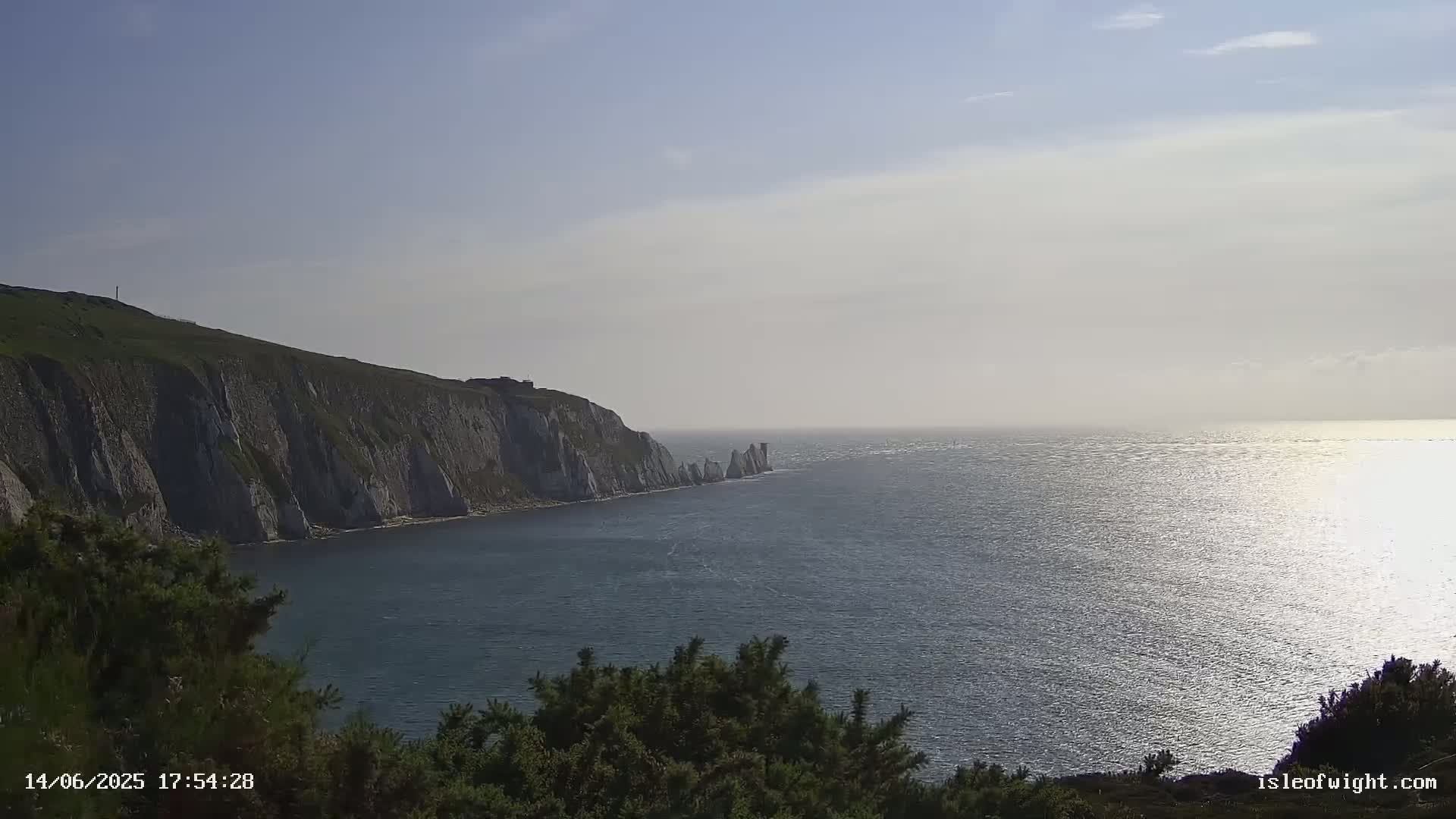 White cliffs rise from a calm, sunlit sea, with a line of smaller rock formations visible further out.