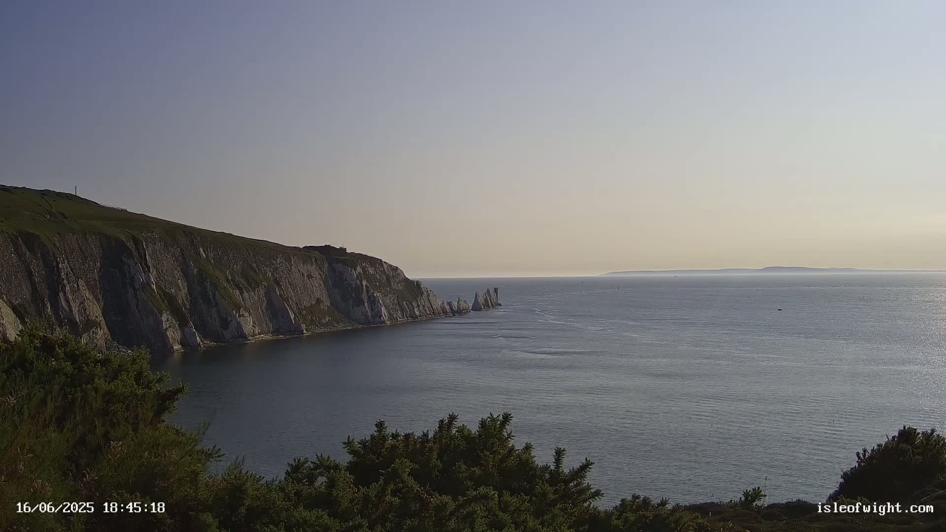 White cliffs rise from a calm sea under a clear, light sky, with a distant landmass visible on the horizon.