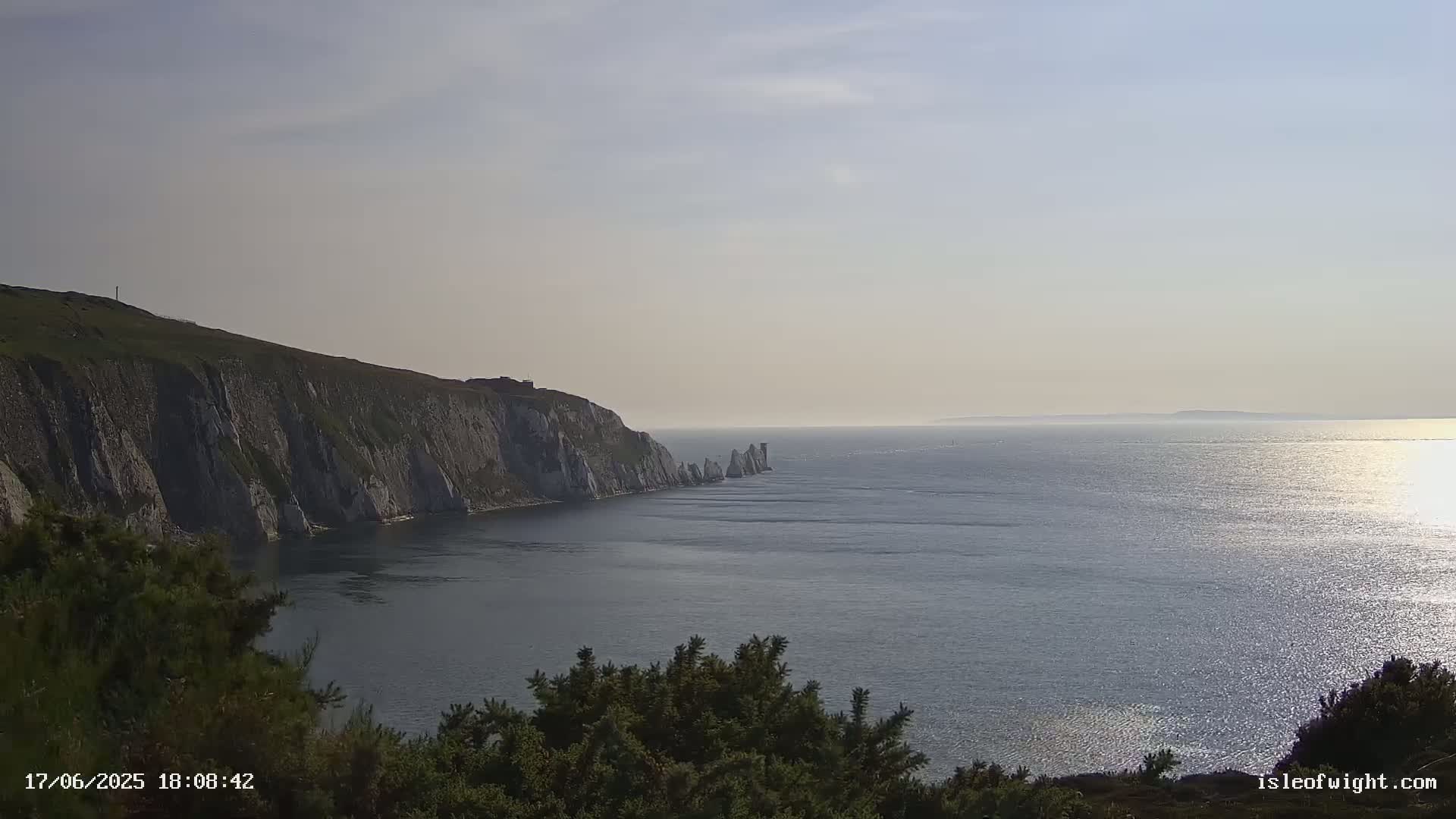 White cliffs extend into a calm, sunlit sea where several tall, thin rock formations rise from the water.