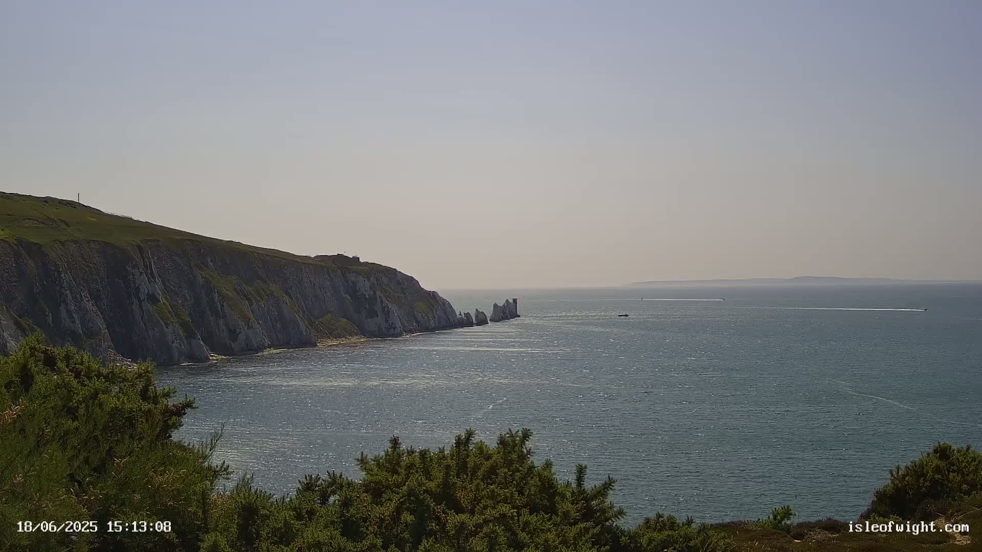 White cliffs meet a calm, blue sea under a hazy sky, with a few boats visible on the horizon.