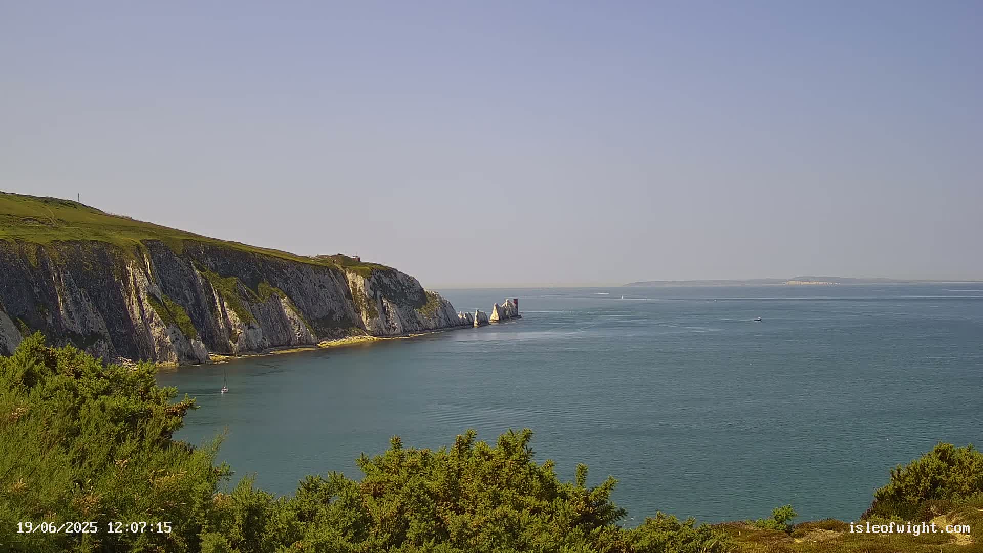 A calm, sunny day reveals a picturesque coastal scene with white cliffs, green hills, and the Needles rocks jutting out into a tranquil blue sea.