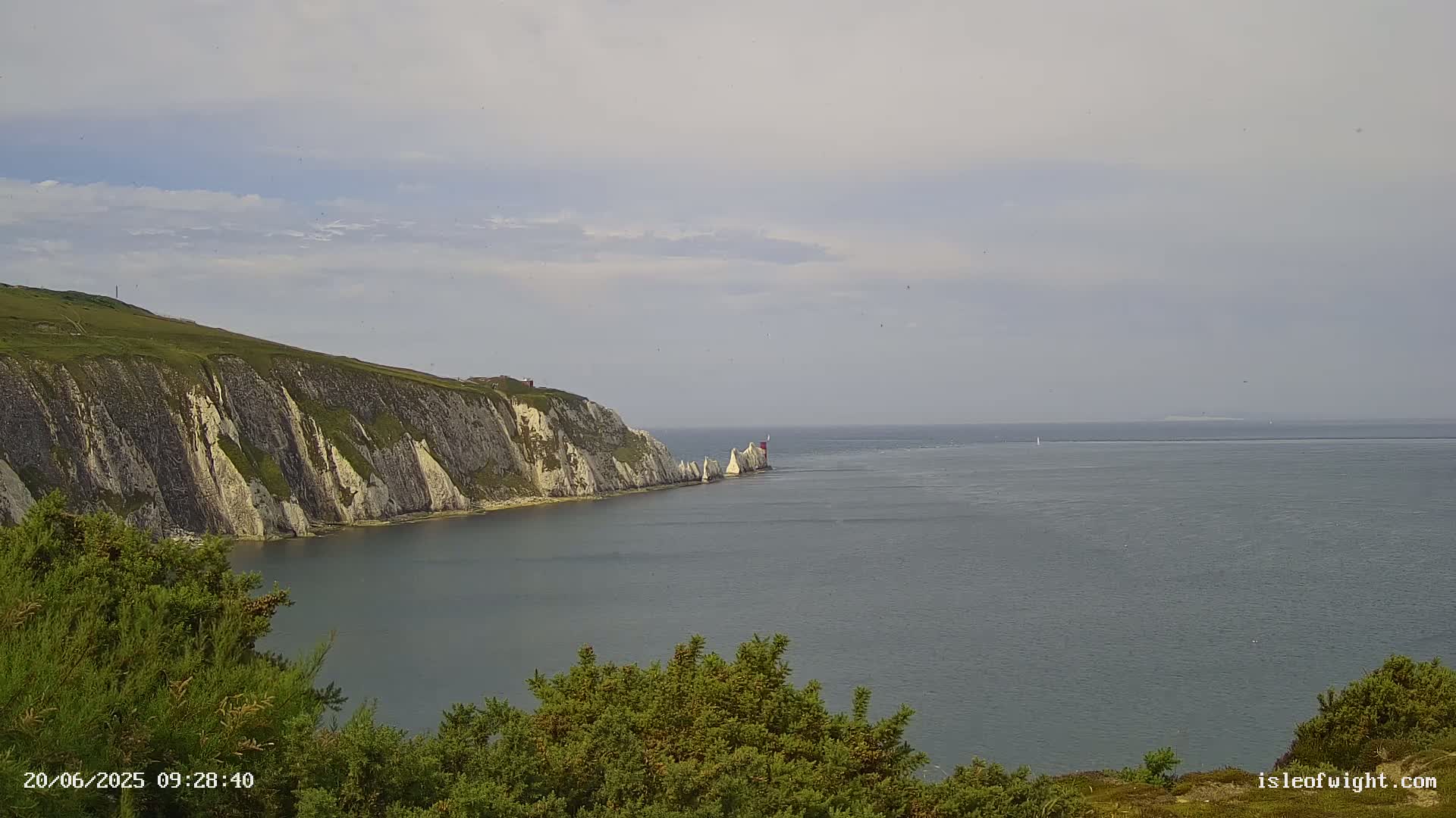 White cliffs and a lighthouse are seen from a distance, overlooking a calm, gray sea under a mostly cloudy sky.