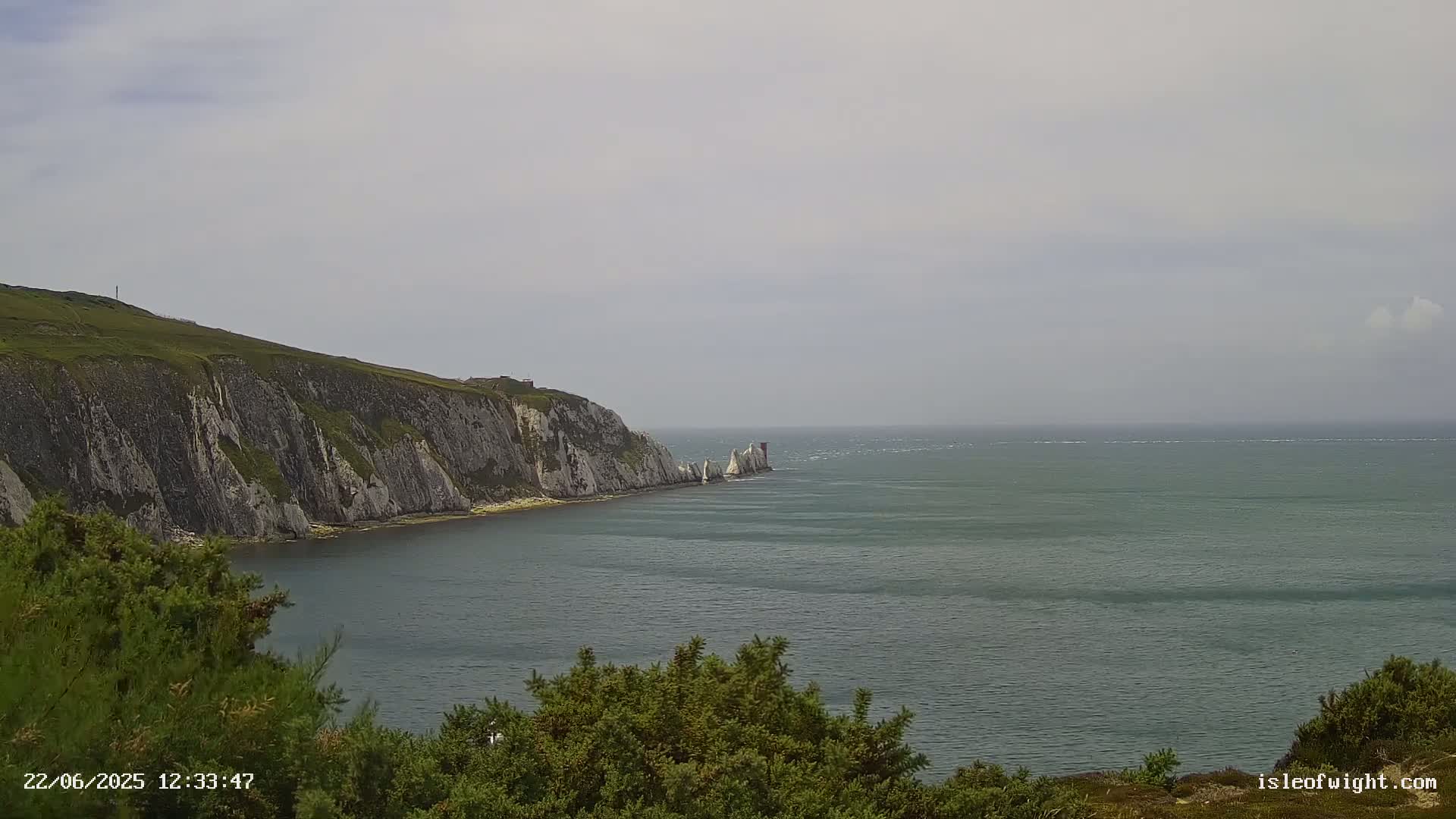 A view of a rocky, green cliff overlooking a calm, grey-green sea with a cluster of white rocks visible in the distance under a mostly cloudy sky.