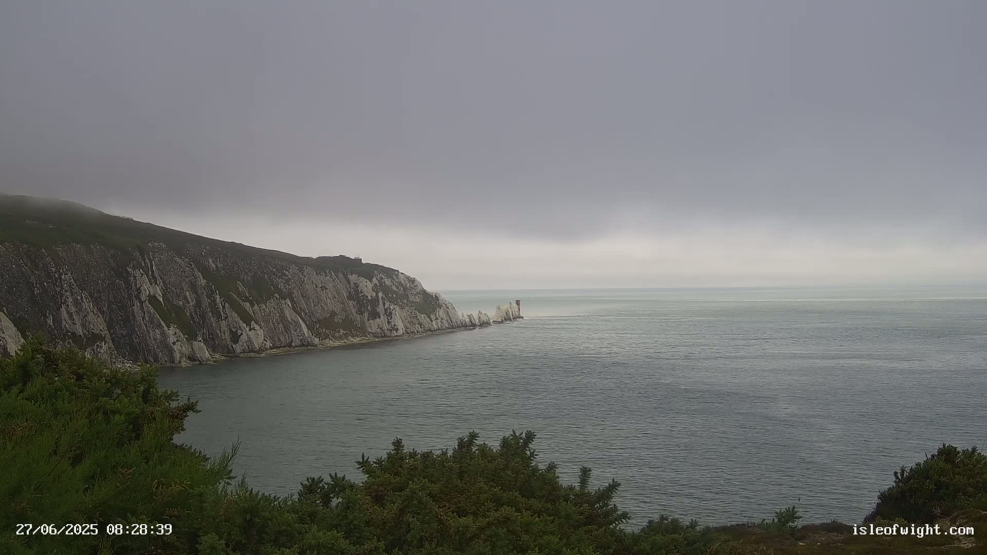 A gray, overcast day reveals a coastal scene with a dramatic white cliff face, partially shrouded in fog, jutting into a calm, gray sea.