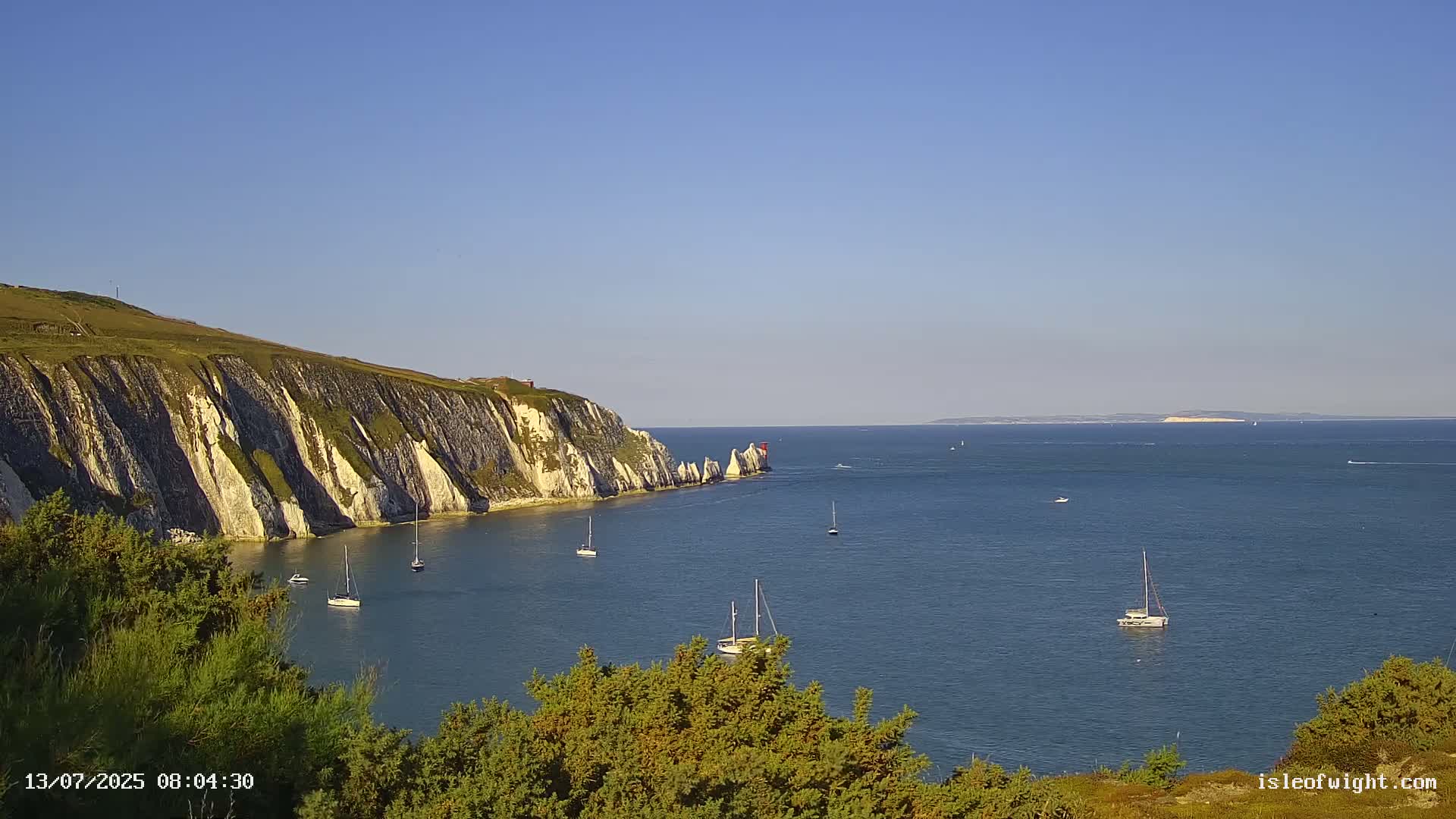 White cliffs rise from a calm, blue sea dotted with sailboats under a clear blue sky.