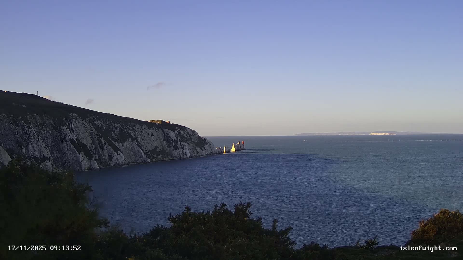 A clear and sunny day showcases a calm blue sea beside white cliffs and the iconic Needles rock formations, capped by a lighthouse, with distant land visible on the horizon.