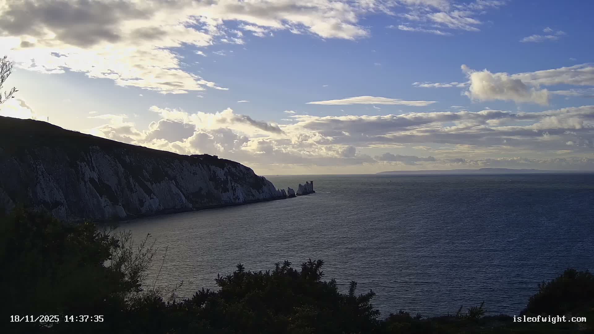 A clear and sunny day showcases a calm blue sea beside white cliffs and the iconic Needles rock formations, capped by a lighthouse, with distant land visible on the horizon.